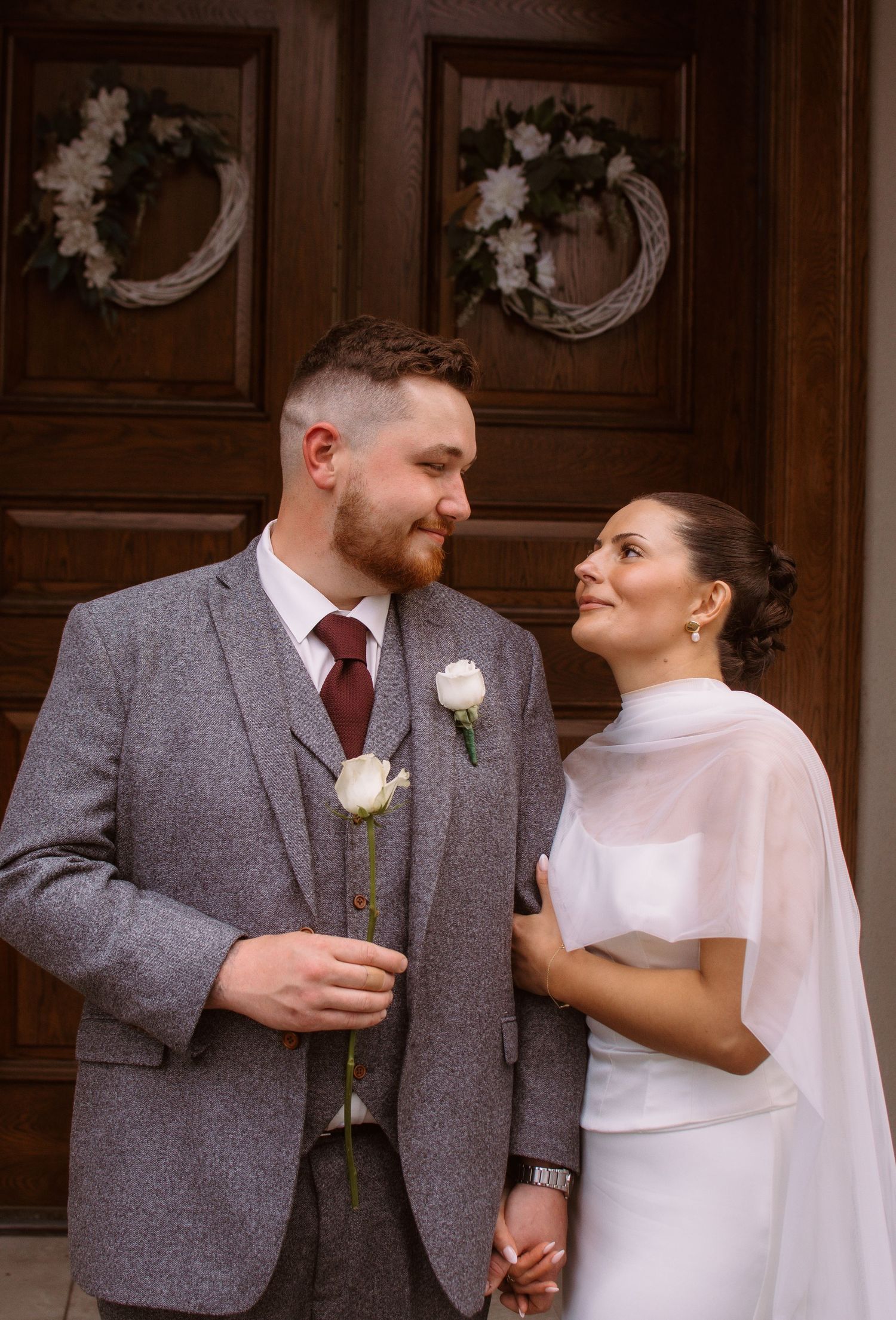A bride and groom smile at each other in wedding attire before a wooden door with wreaths.