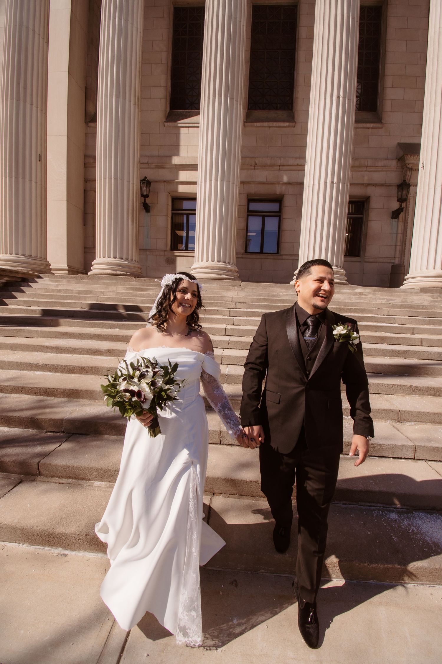 Bride in white gown and groom in black suit hold hands walking down steps of columned building on wedding day.