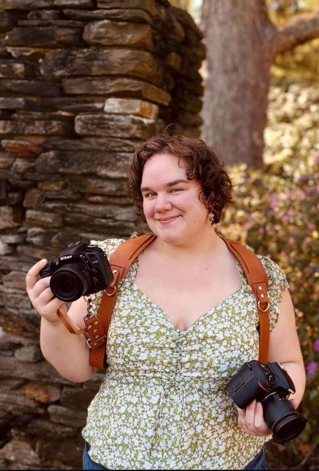 A smiling woman holds two Nikon cameras outdoors near a rustic stone wall, wearing a floral top with a brown leather strap.