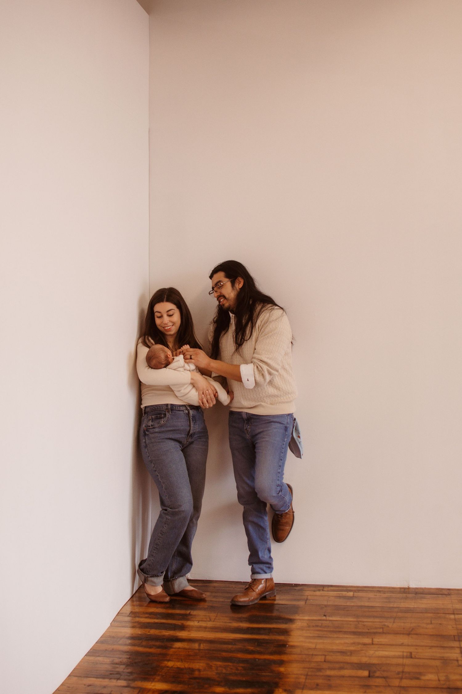 A couple leans against a white wall on hardwood floors, lovingly gazing at their newborn baby wrapped in a blanket.