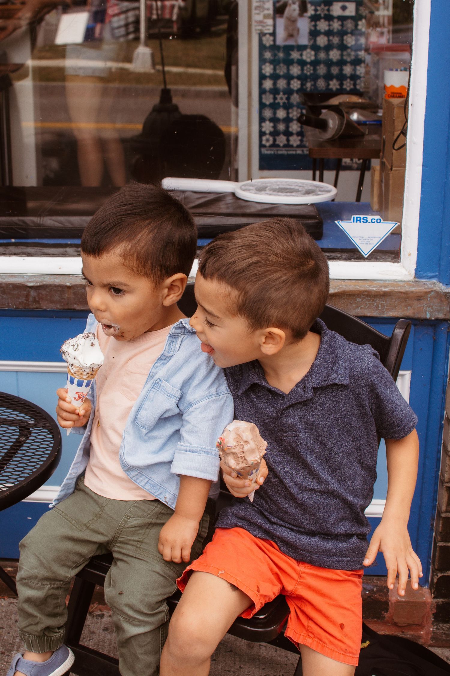 Two young boys sitting together eating ice cream cones outside an ice cream shop.