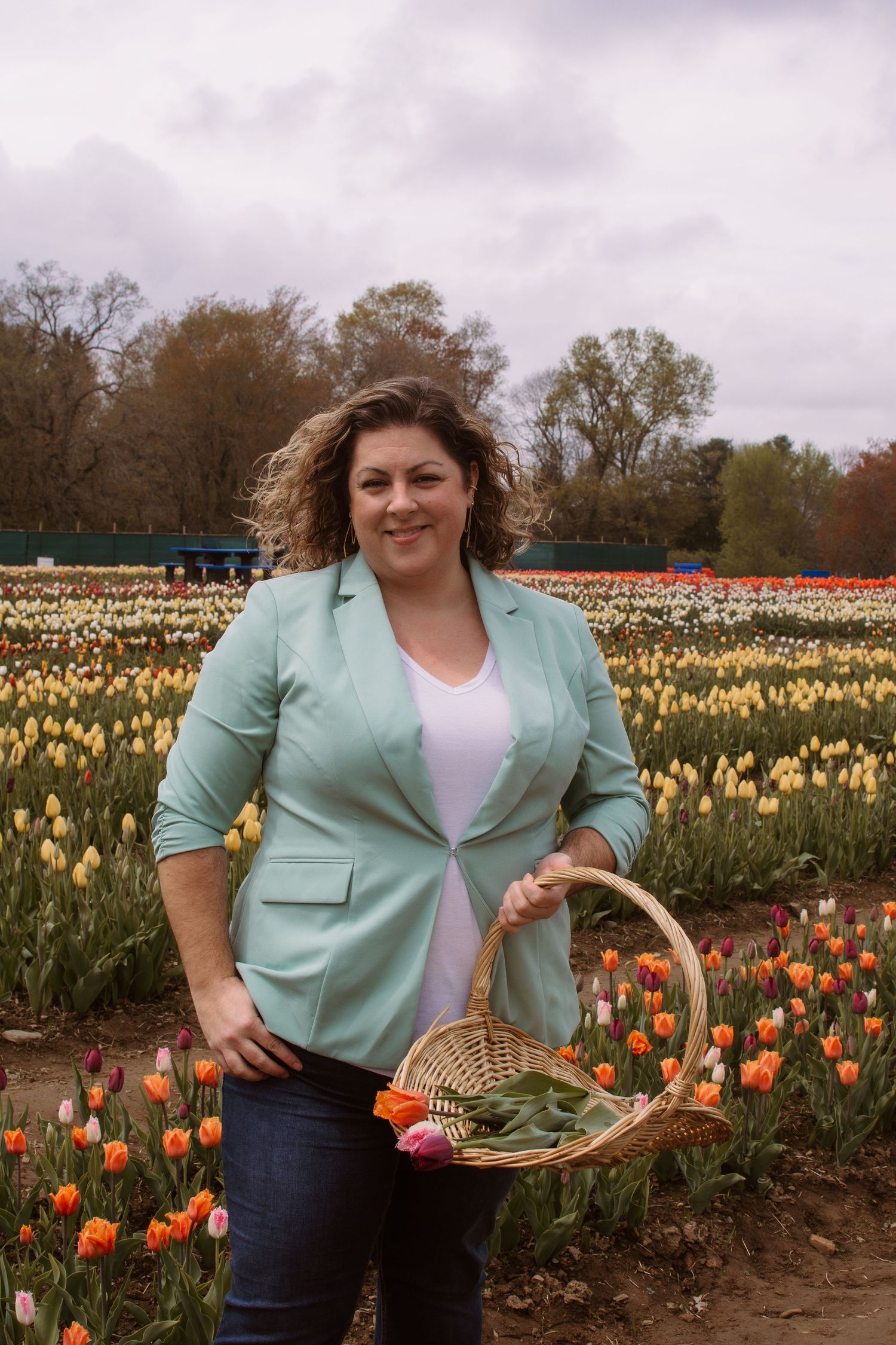 Woman in mint blazer holding wicker basket of tulips, smiling in colorful tulip field on cloudy day.