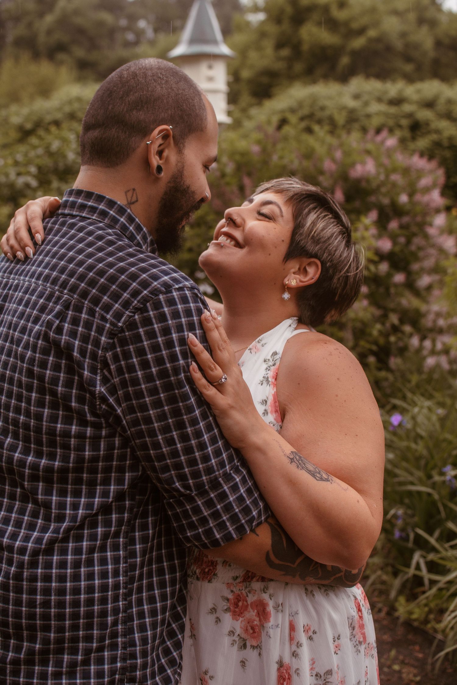 Couple laughing and embracing outdoors in a garden, woman in floral dress with tattoos, man in plaid shirt.