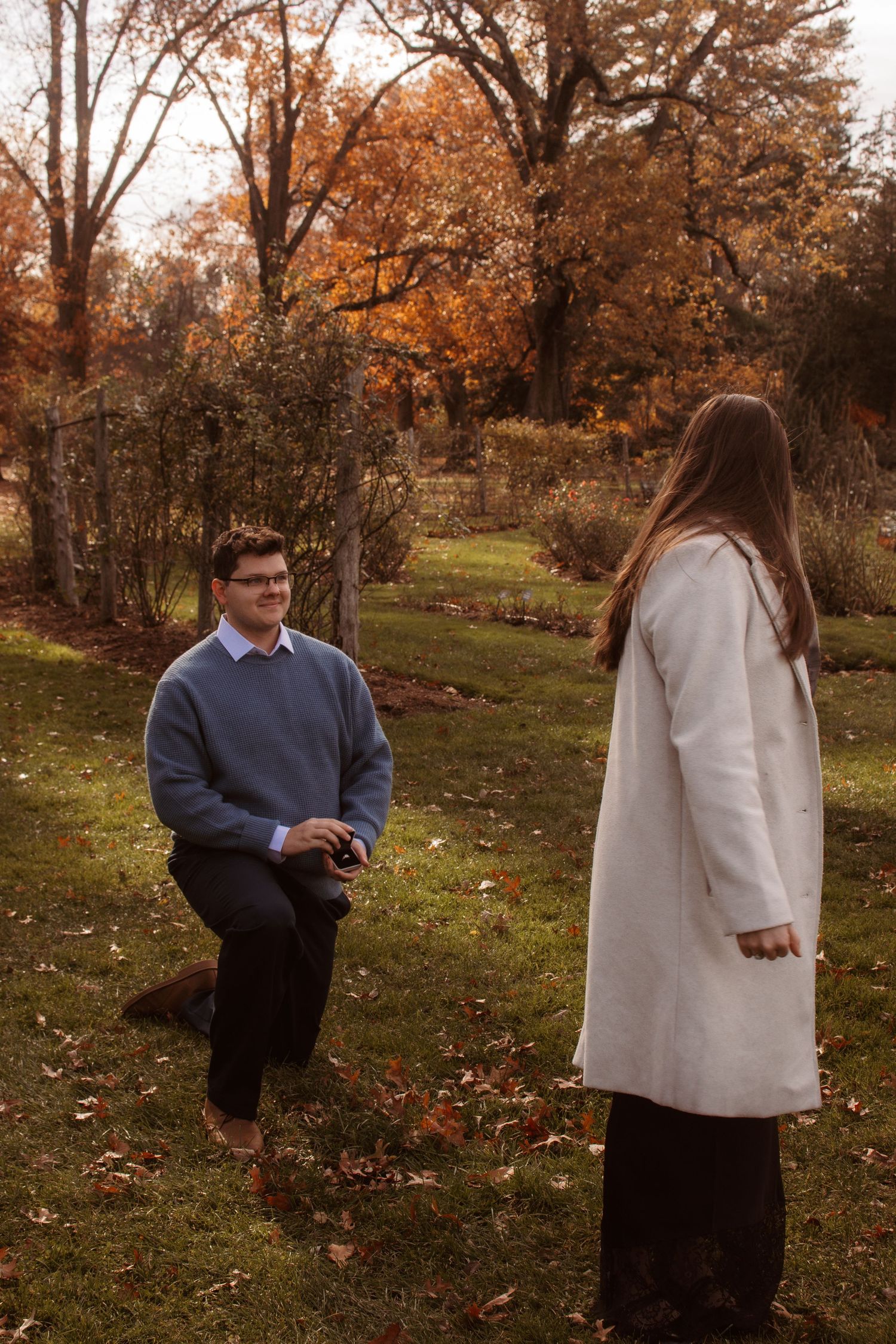 A couple in autumn attire playfully interacts in a park setting with fallen leaves and orange trees.