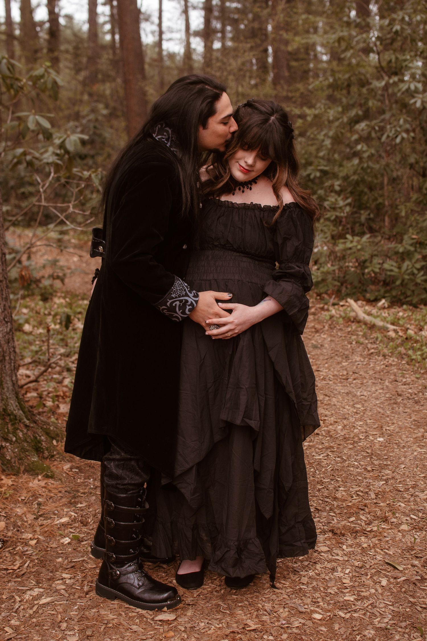 Gothic couple in black medieval attire embracing on forest path, pregnant woman cradled tenderly by partner.