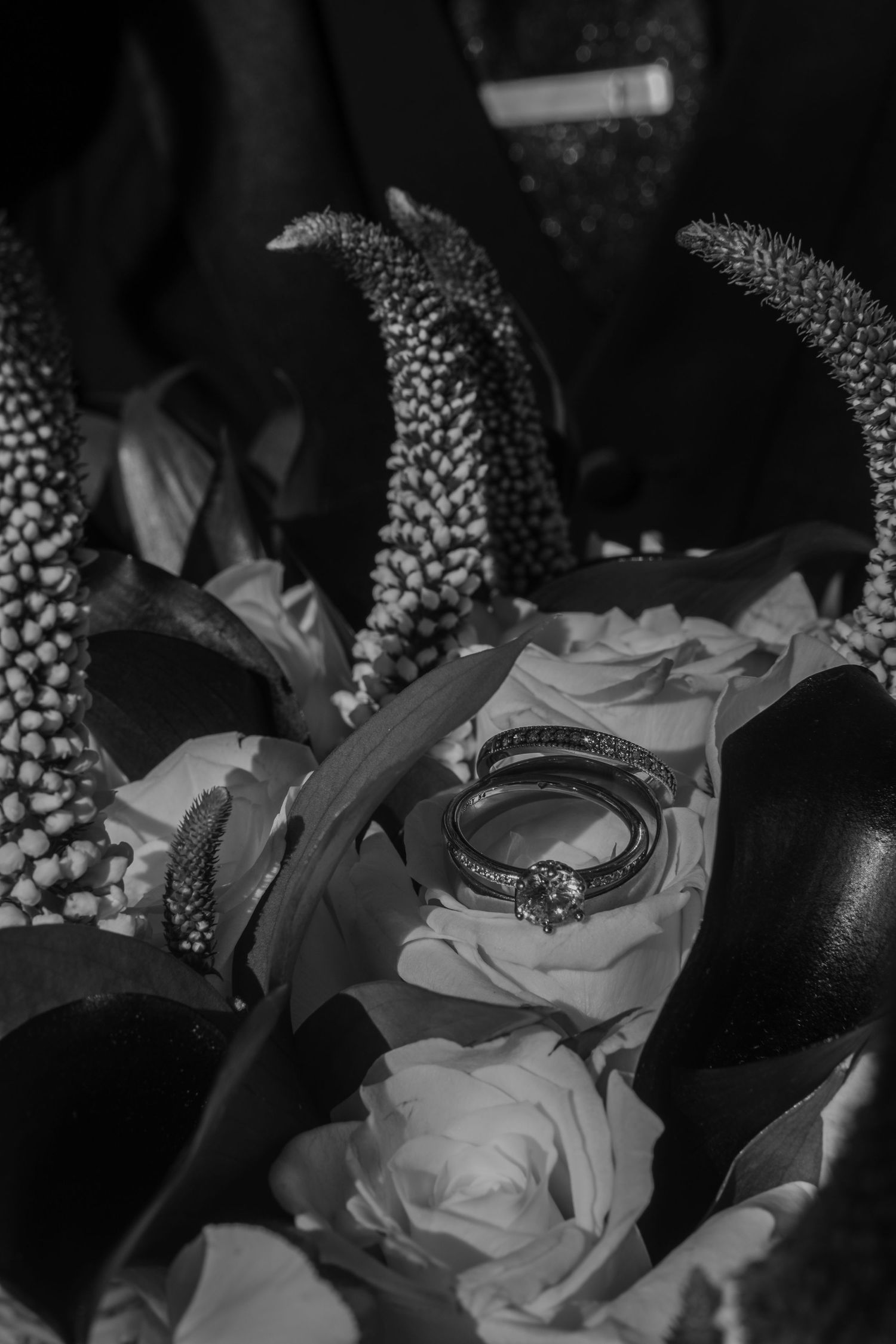 Black and white photo of wedding rings resting on roses and exotic flowers in a bridal bouquet arrangement.