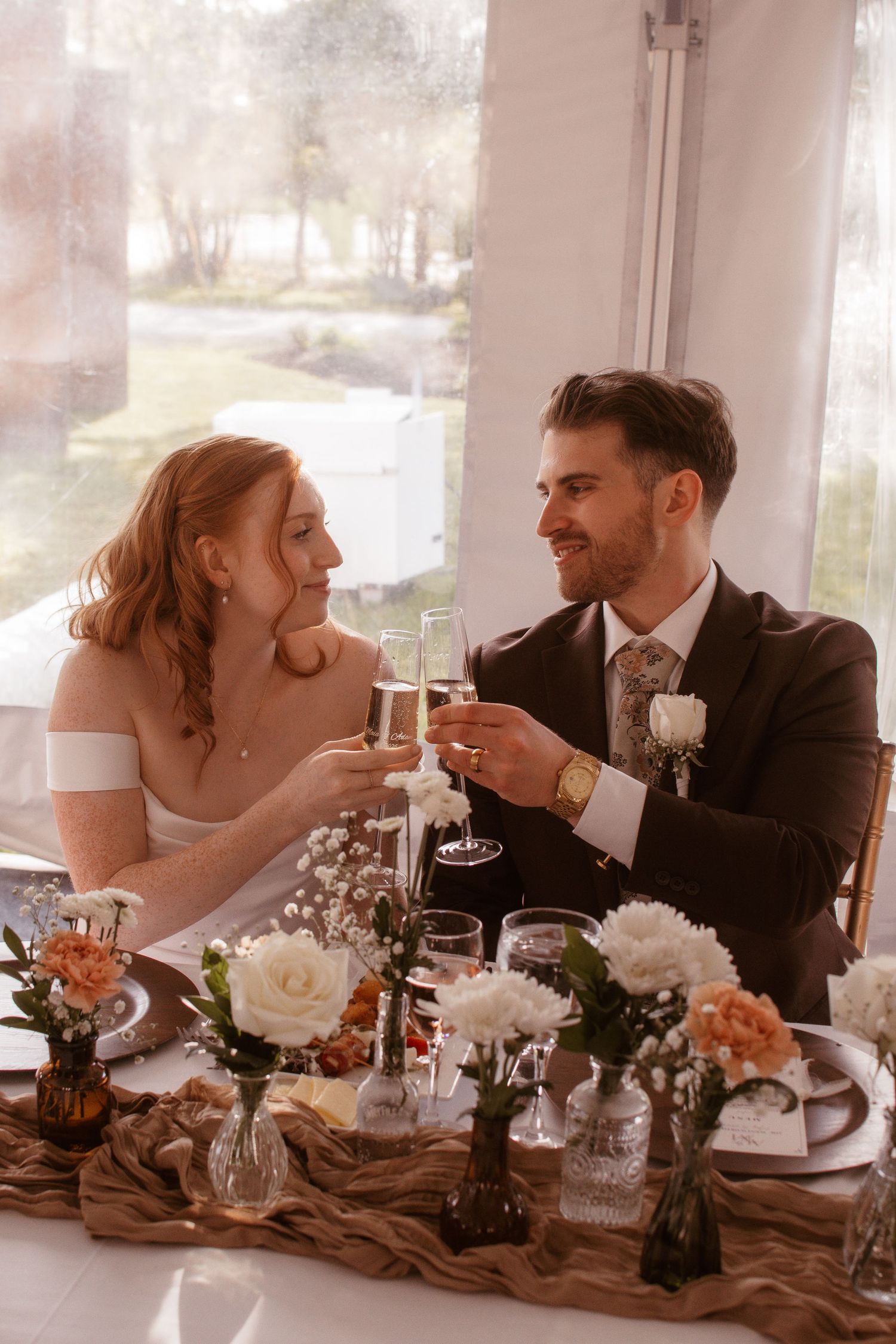 Bride and groom toasting with champagne glasses at floral-decorated sweetheart table during wedding reception.