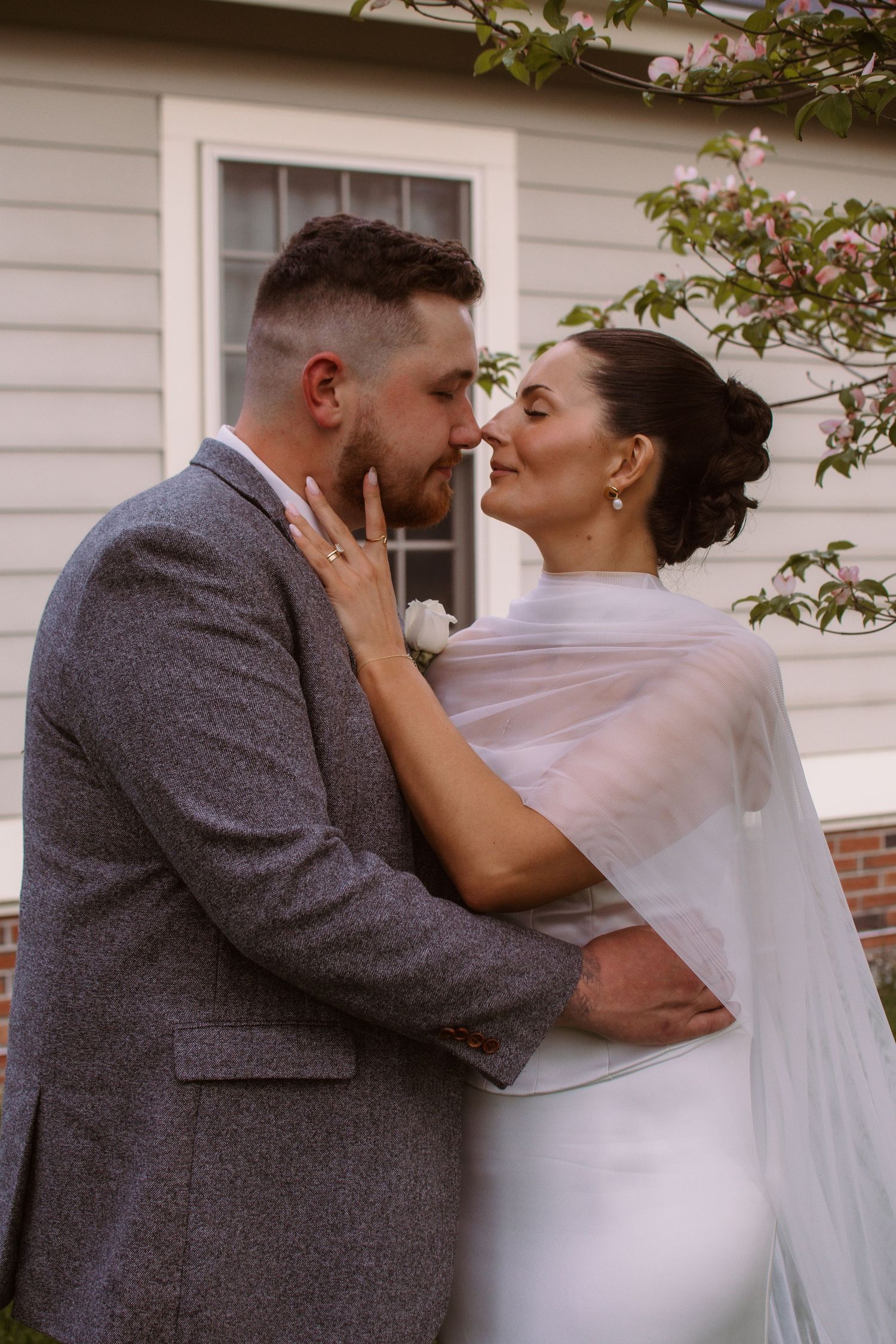 A bride and groom share a romantic almost-kiss outdoors, with blooming trees and a house in the background.