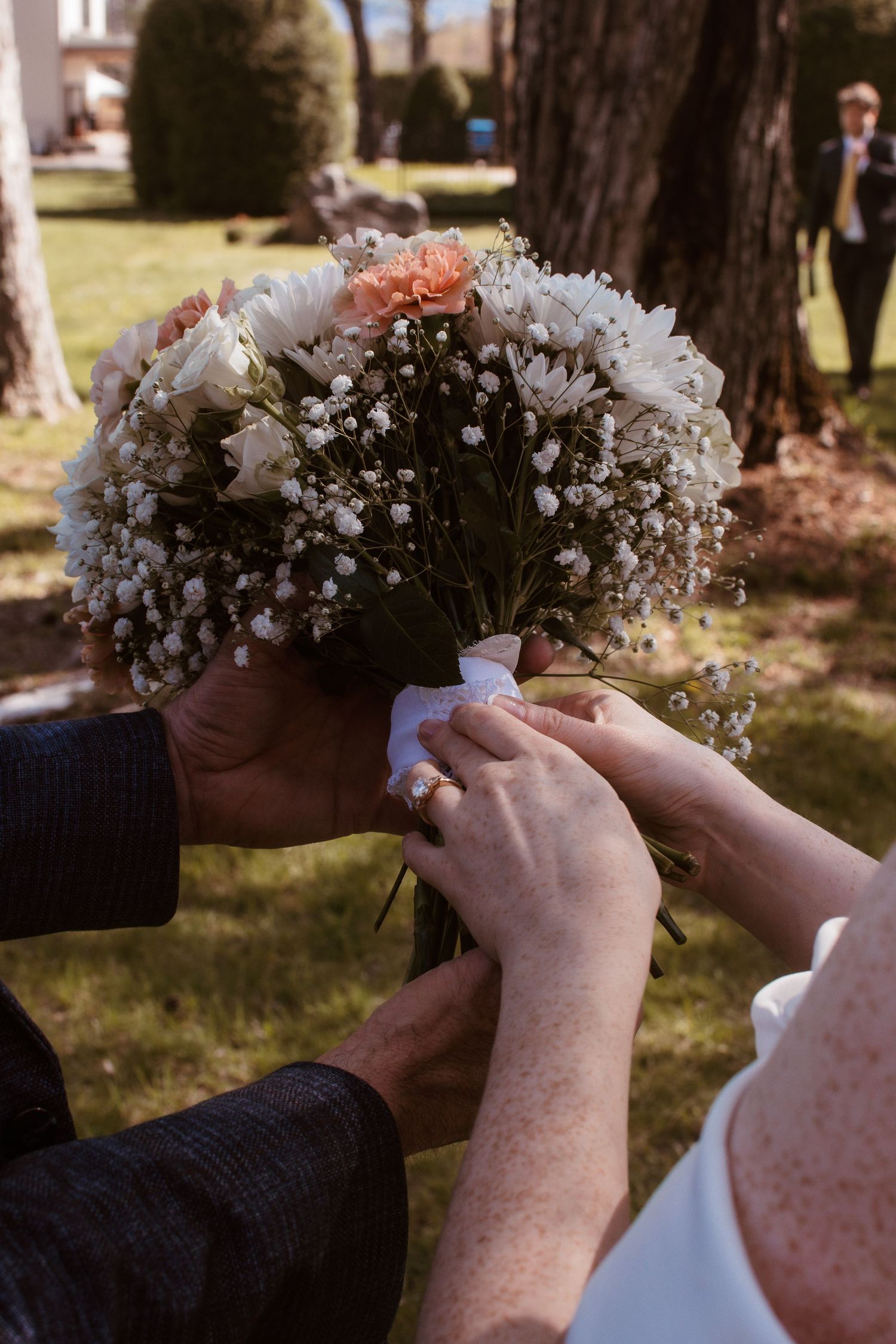 Hands holding a wedding bouquet of white chrysanthemums, baby's breath, and peach roses tied with white ribbon outdoors.