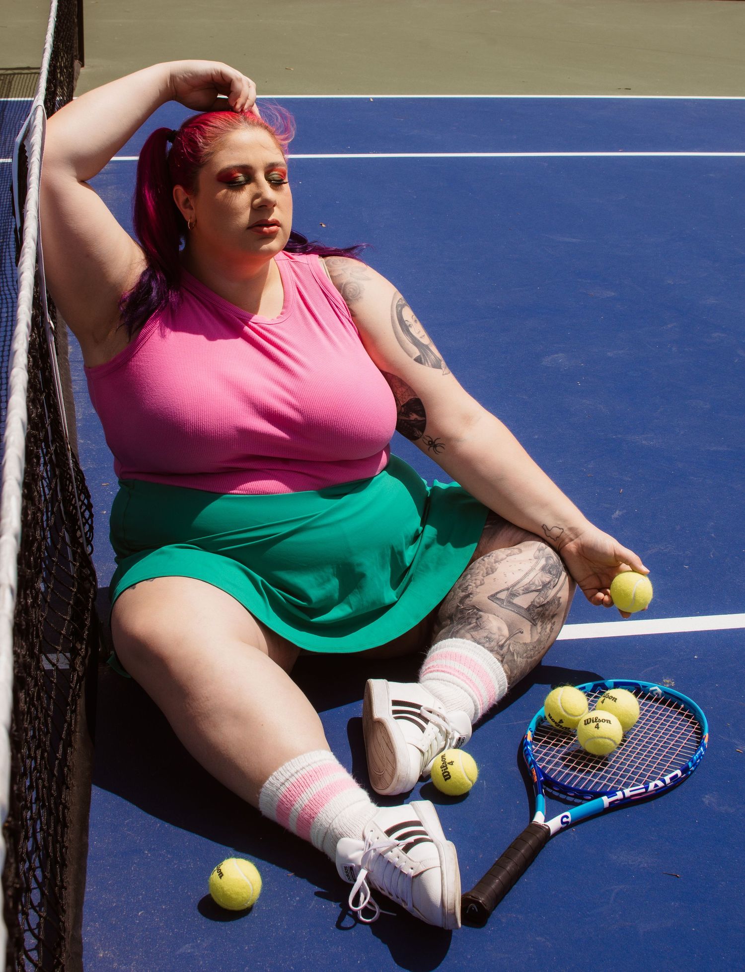 A person in pink and green sportswear sits on a tennis court with tennis balls and racquet nearby.