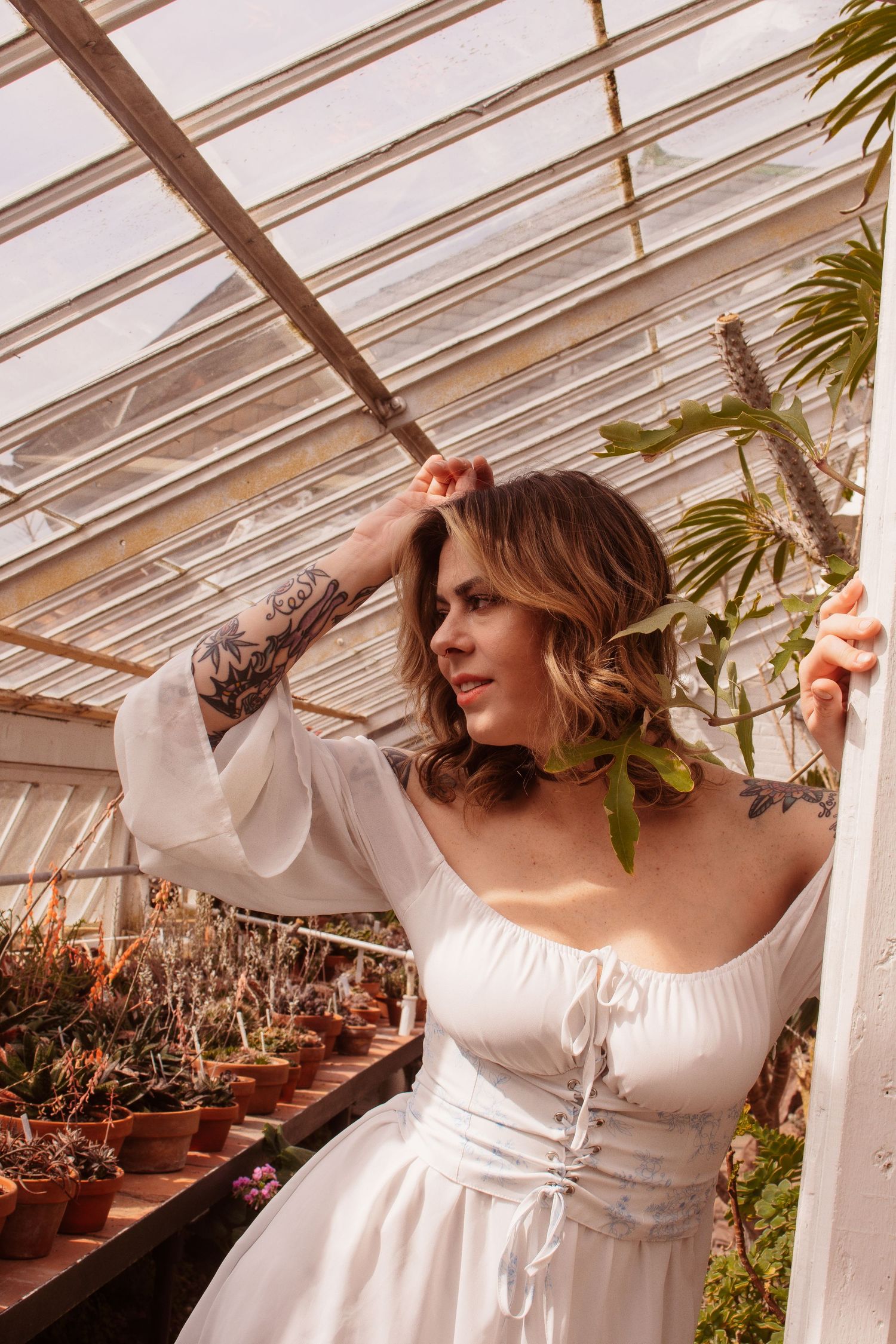 Woman with tattoos in white dress poses gracefully in a sunlit greenhouse surrounded by potted plants and cacti.