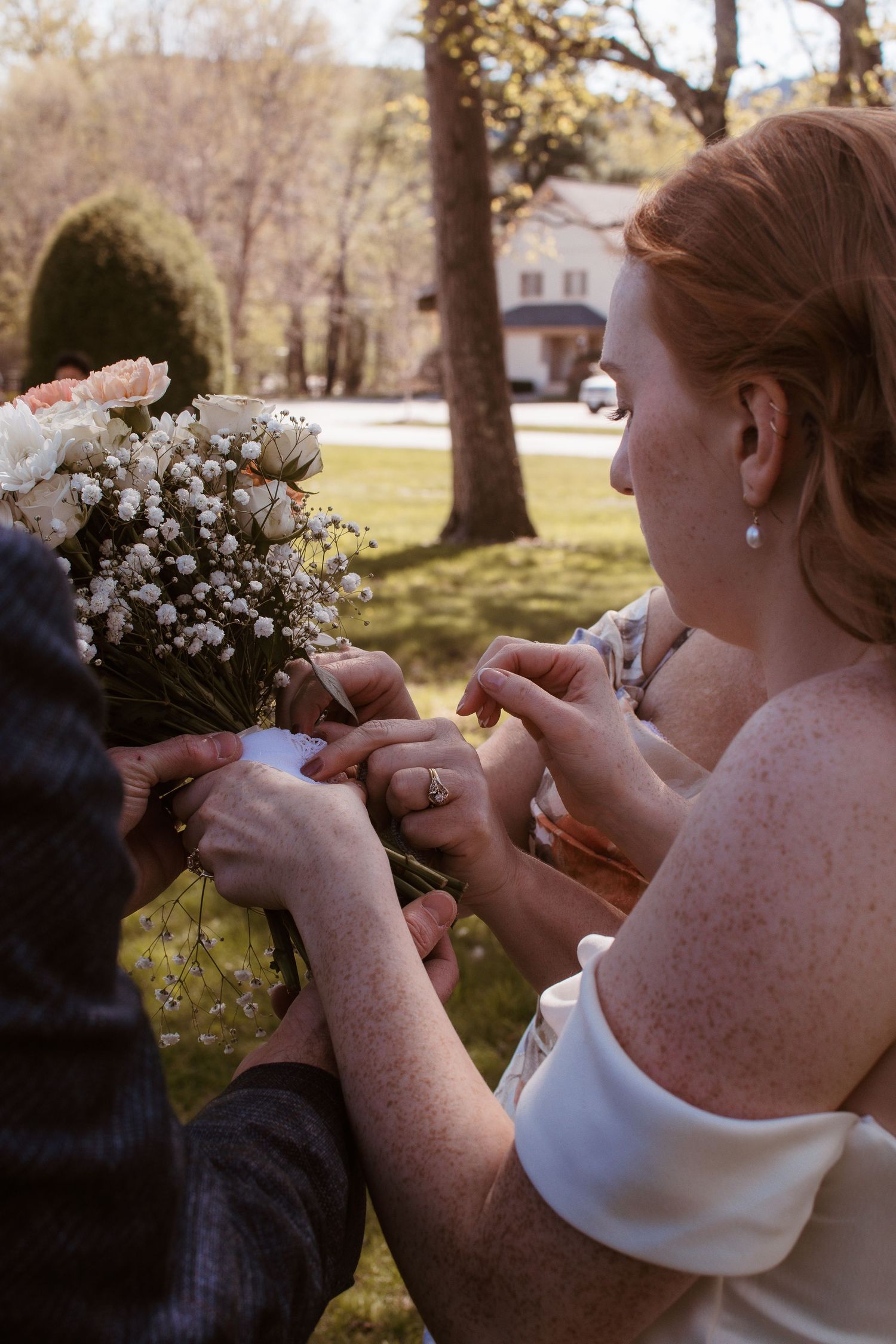 Red-haired bride in white dress arranging a white floral bouquet outdoors at a wedding ceremony.