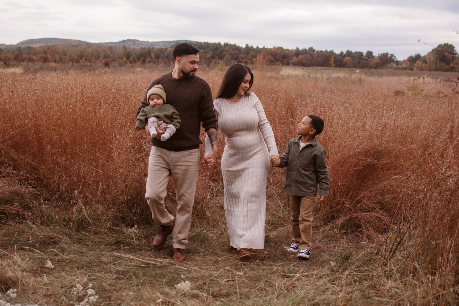 Family of four walking through autumn field, parents holding children's hands, tall brown grass, overcast sky.