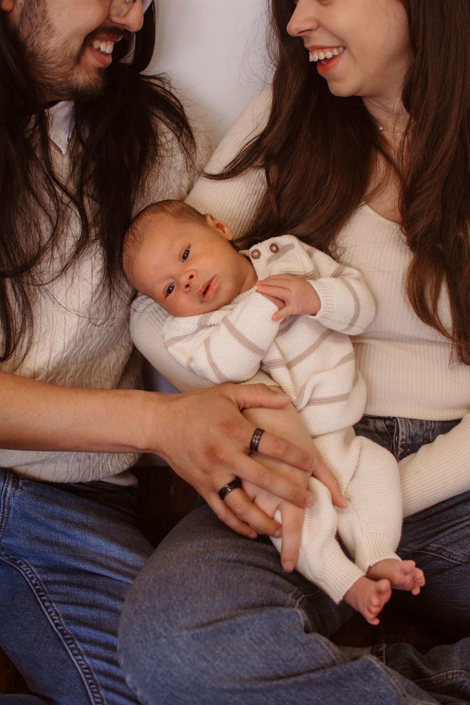 New parents smiling and cradling their newborn baby dressed in a white striped onesie while sitting together.