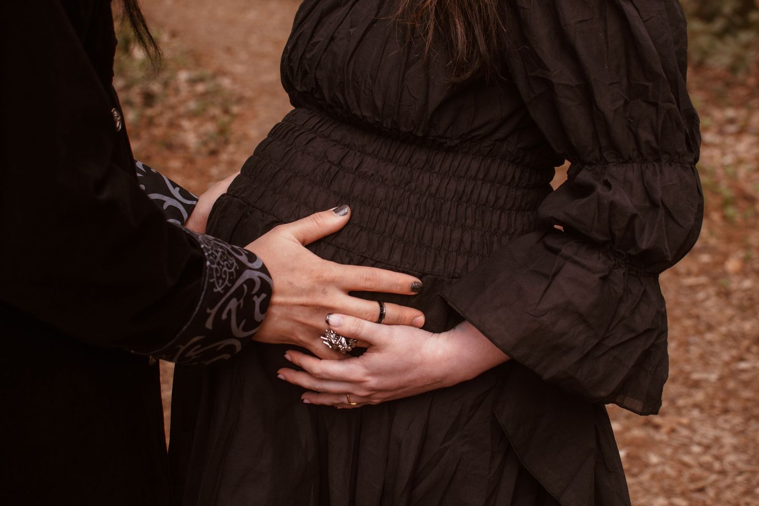 Pregnant woman in black dress with partner's hands cradling baby bump during maternity photo session outdoors.