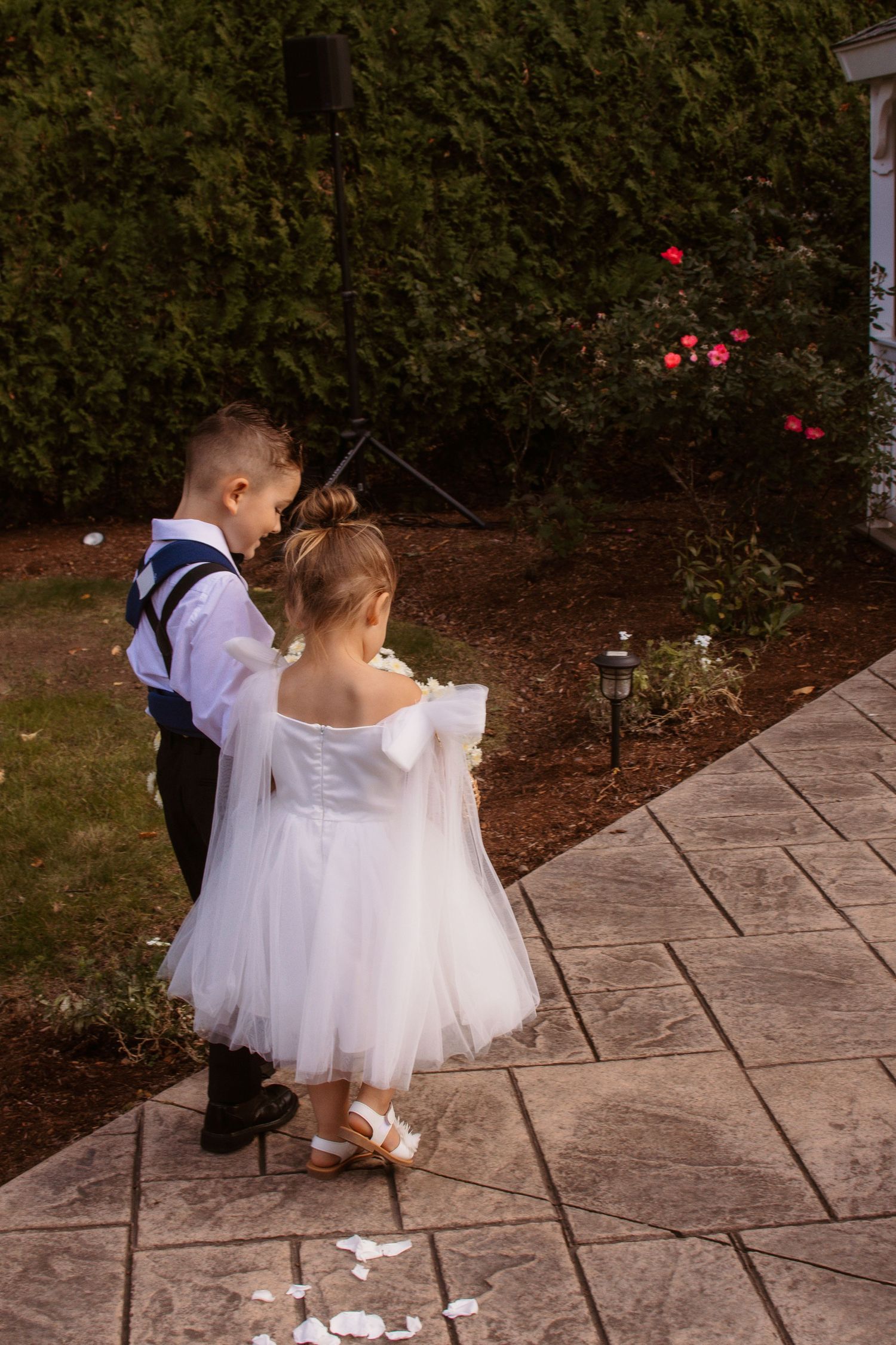 Two young children in formal attire stand together on a stone pathway near flowering bushes at a wedding celebration.