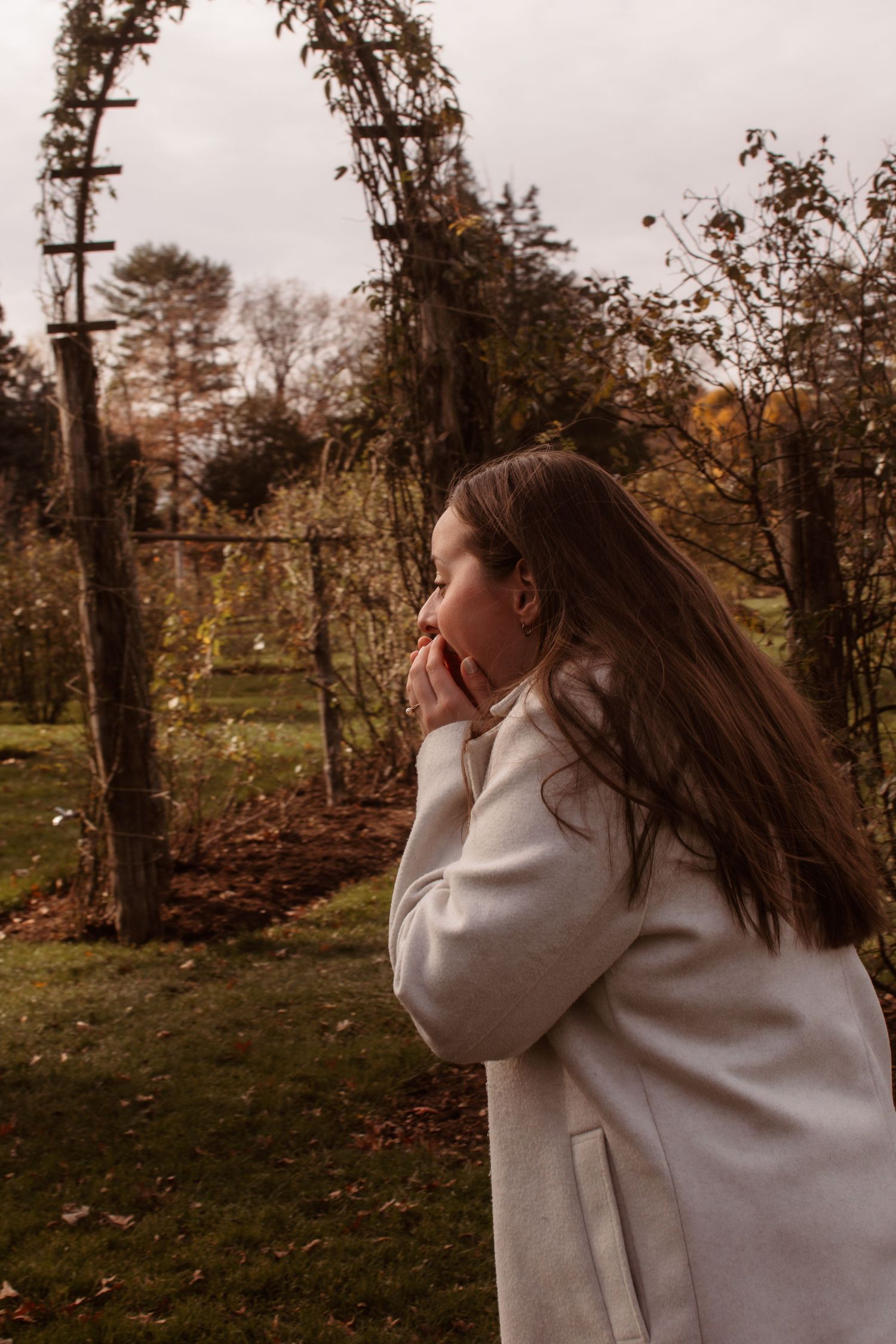 A person in a beige sweater stands in an autumn garden with long flowing hair and trees in the background.