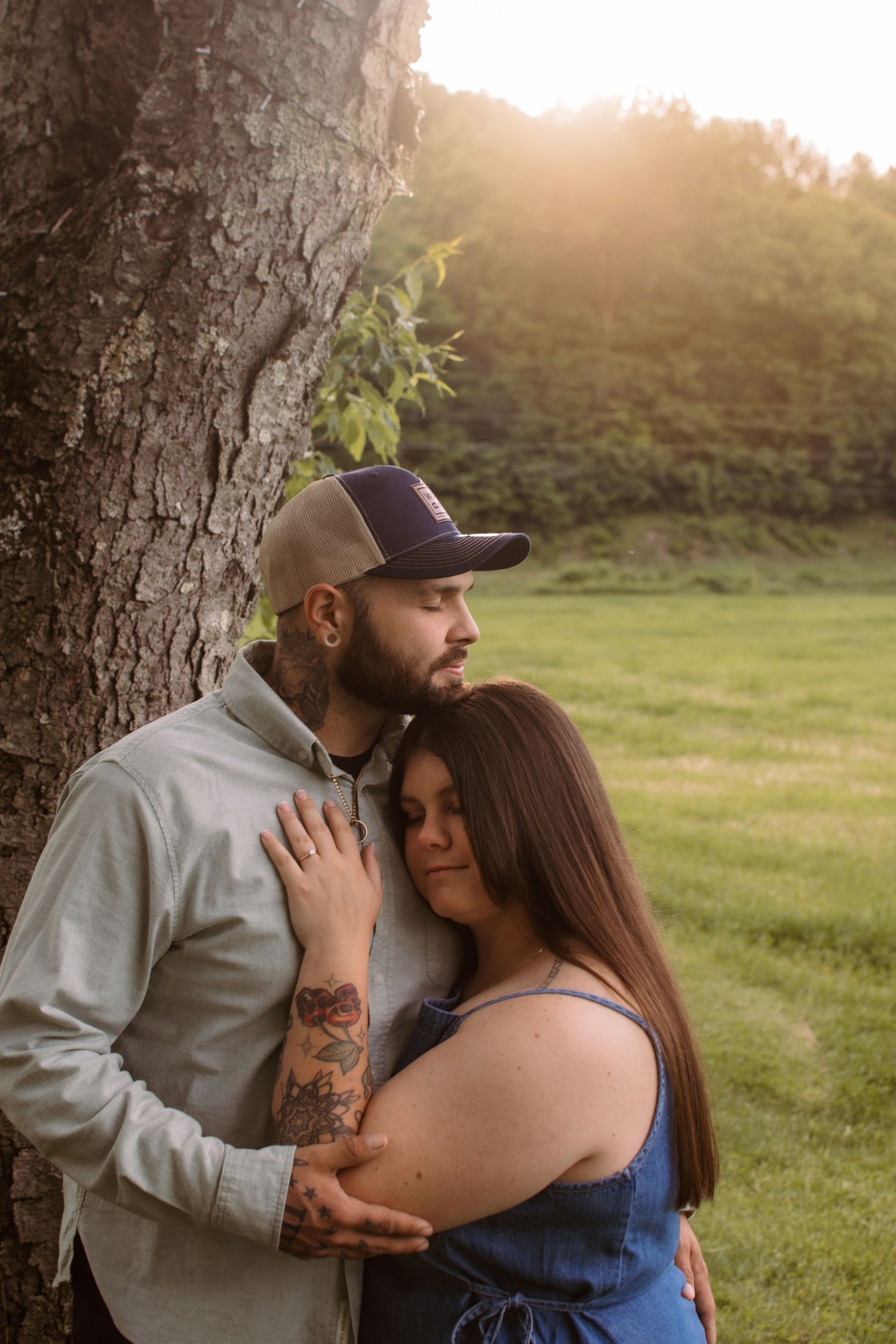 Couple embracing by a tree at golden hour, man with tattoos and cap holding woman in blue dress outdoors.