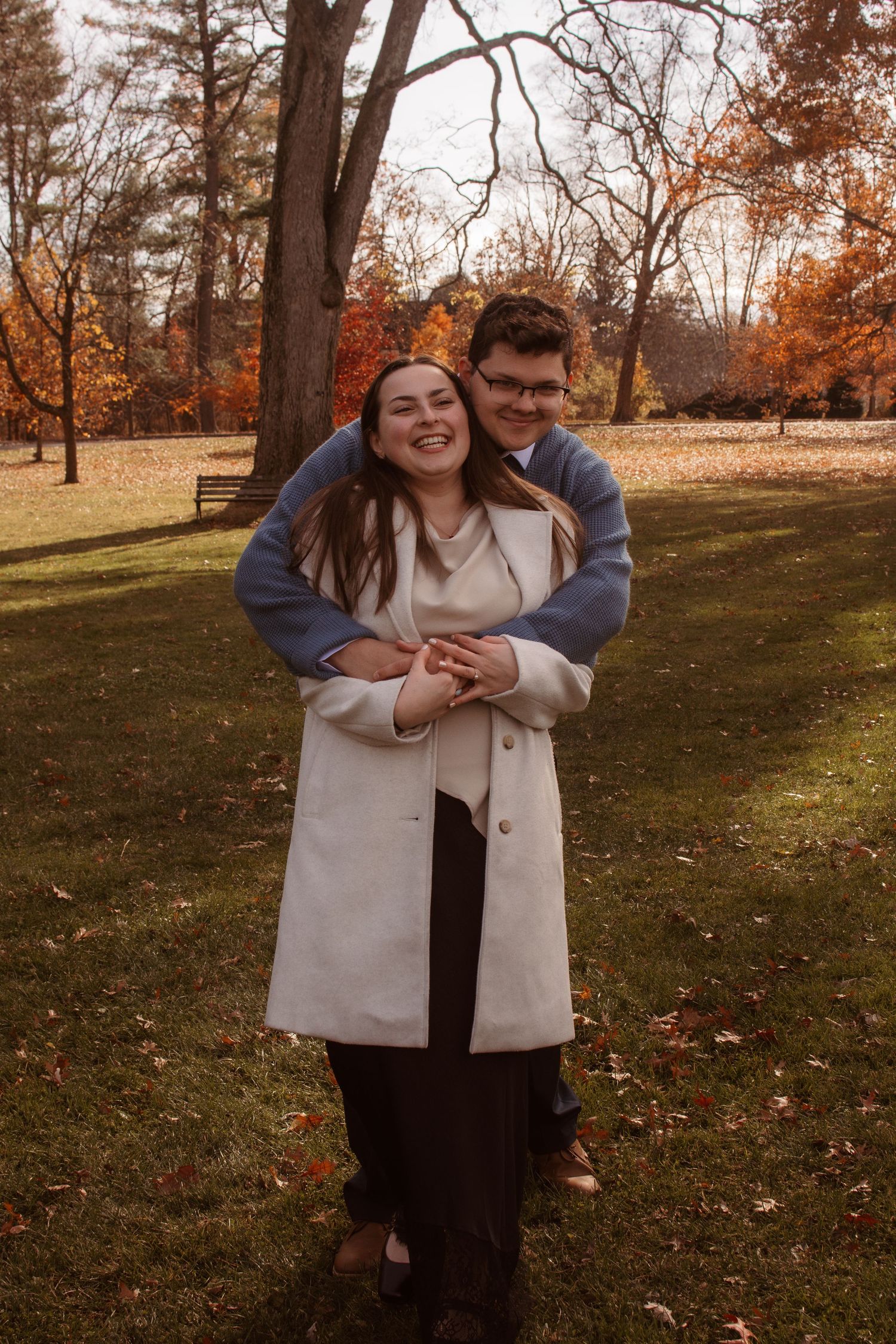 A couple shares a tender moment outdoors on an autumn day with fallen leaves and trees in the background.