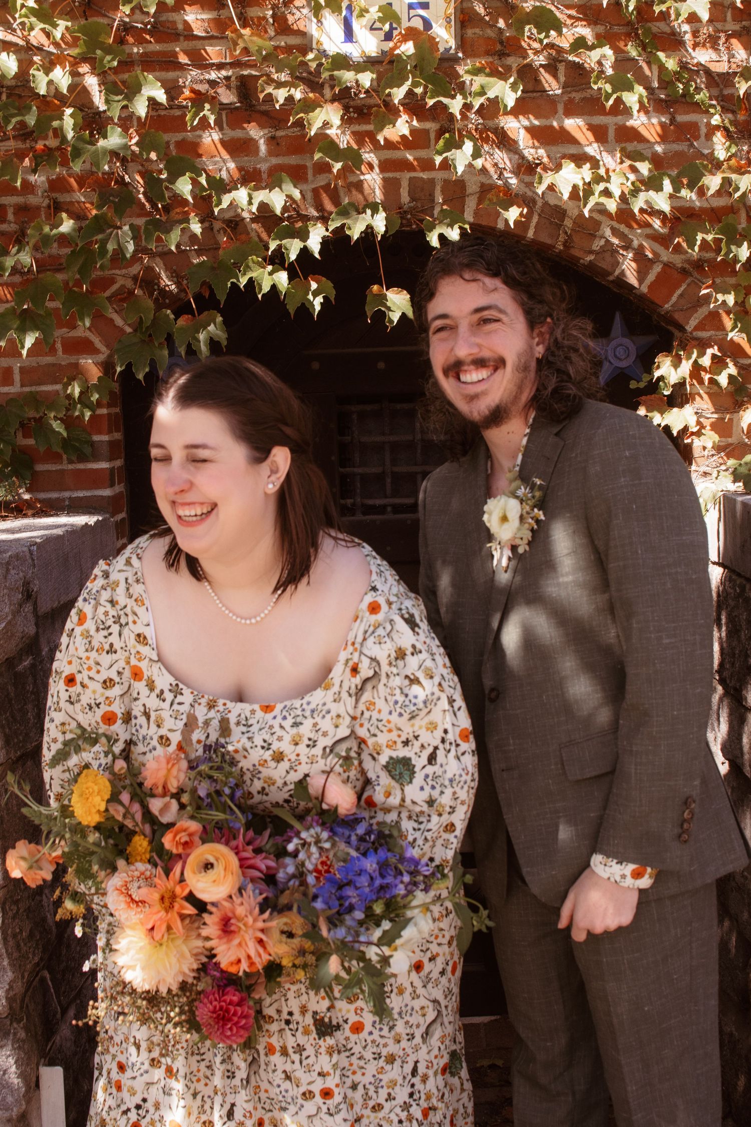 A couple shares a candid moment of laughter while posing with a vibrant autumn bouquet against a brick wall.
