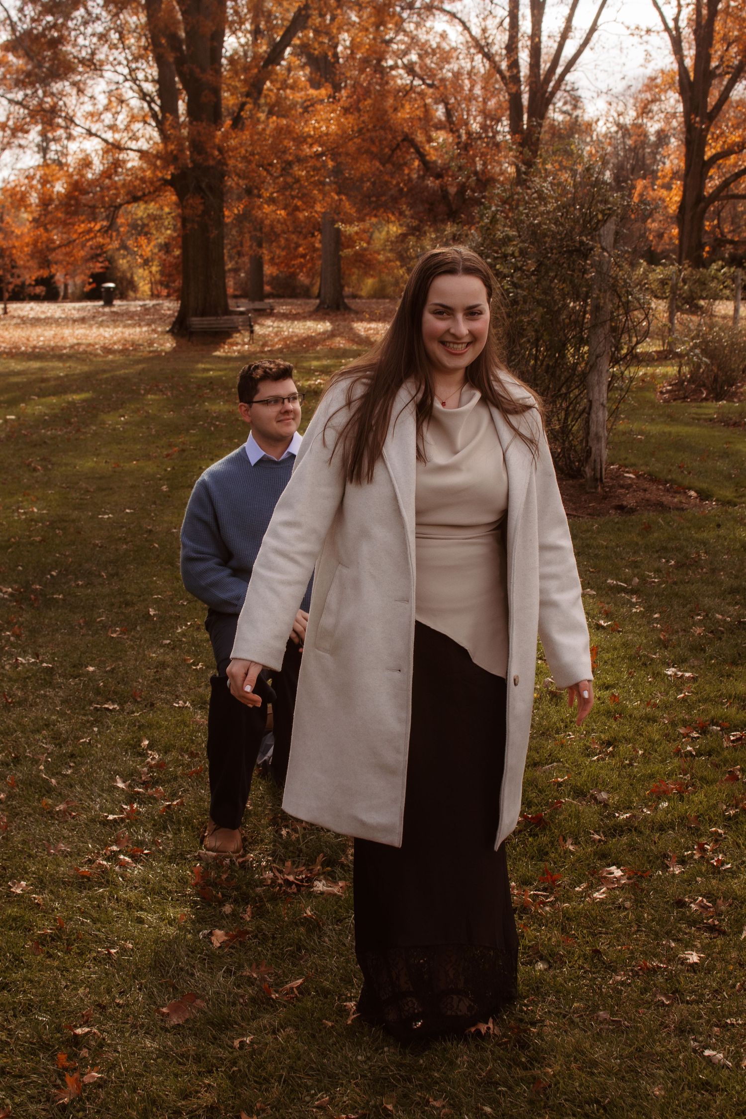 Two people standing outdoors in autumn with fall foliage and trees, wearing coats on grass covered ground.