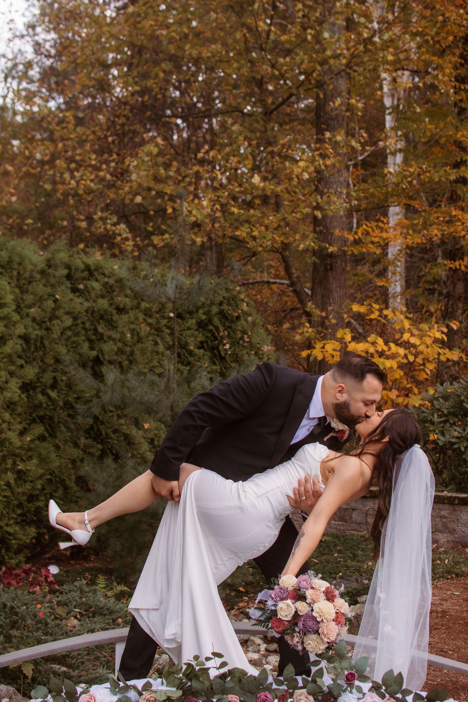 A romantic wedding couple shares a dramatic dip kiss surrounded by autumn foliage and holding a pink floral bouquet.