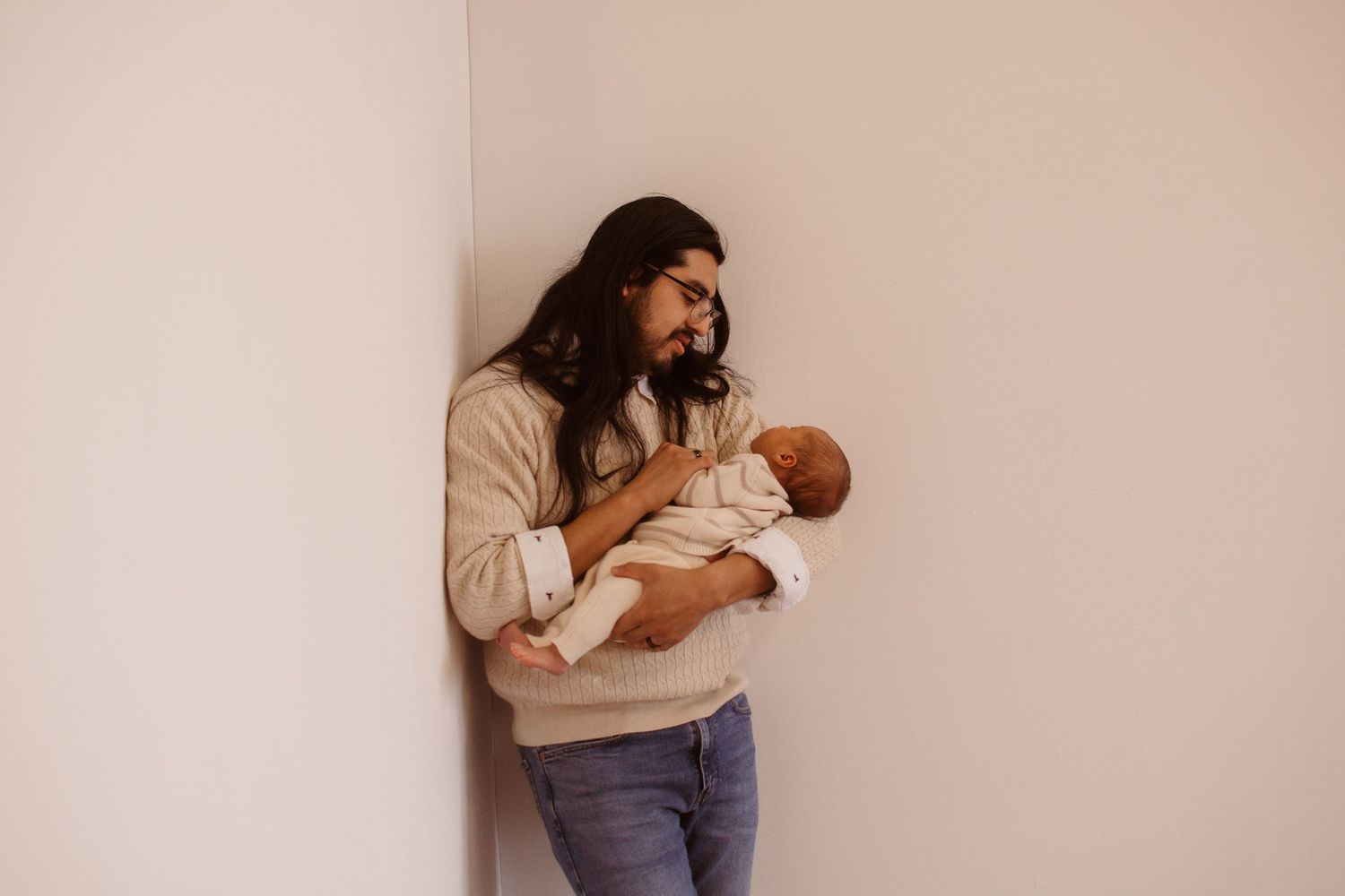 A father with long dark hair and glasses tenderly holds his newborn baby against a white wall.