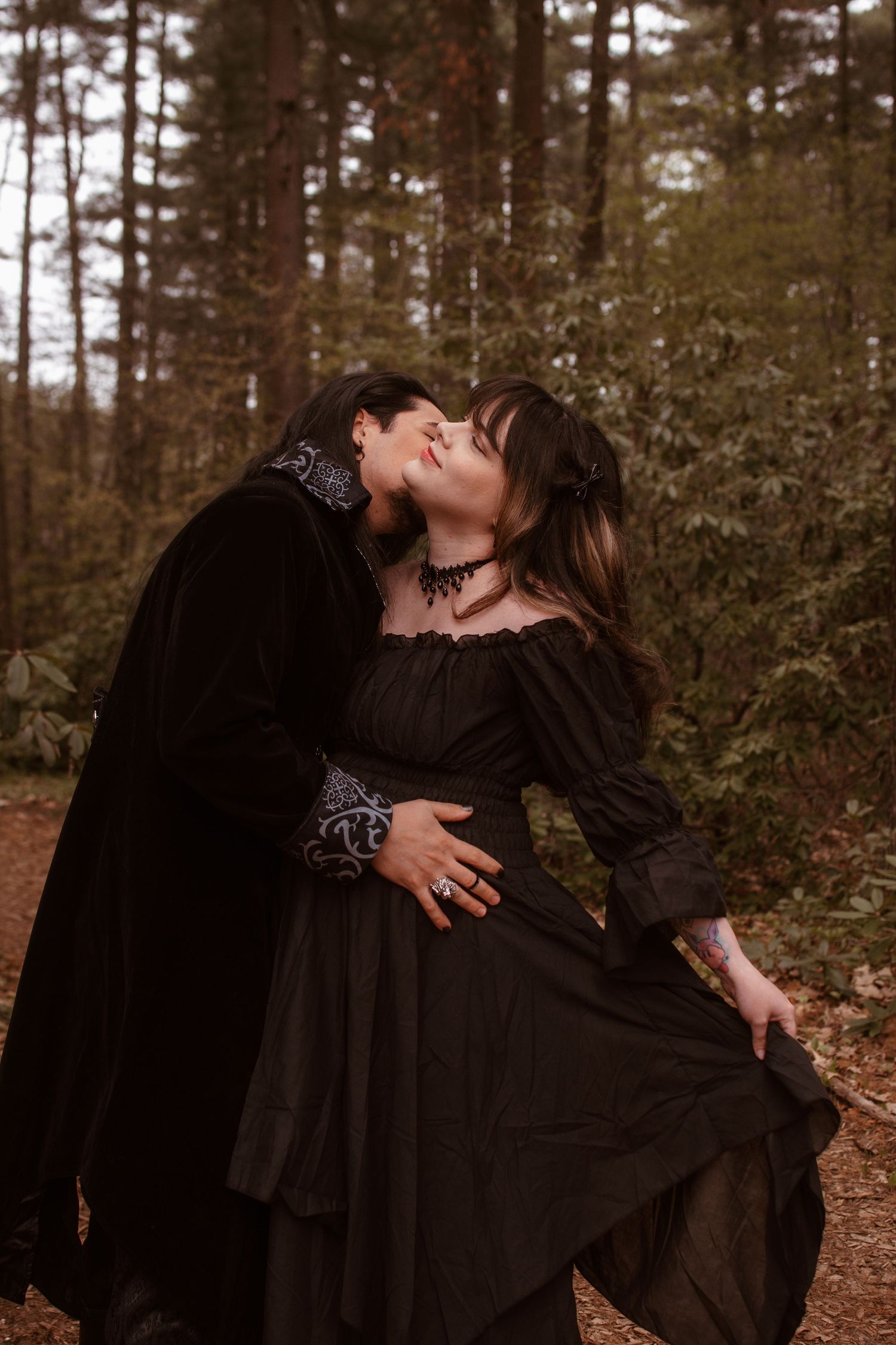 Couple in gothic black Victorian costumes sharing a romantic kiss in a misty forest setting.