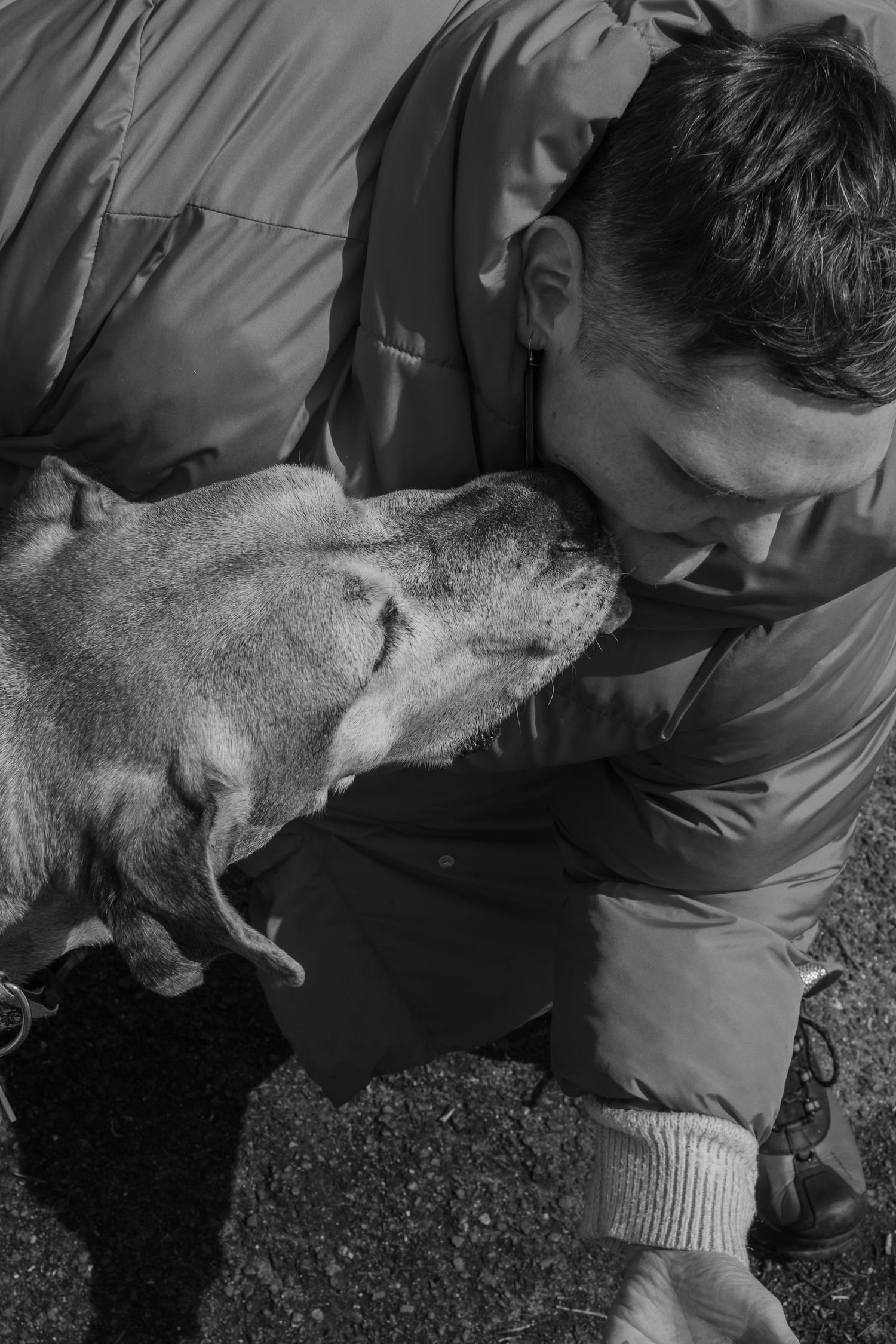 Black and white photo of a man in a jacket affectionately nuzzling with his dog outdoors.