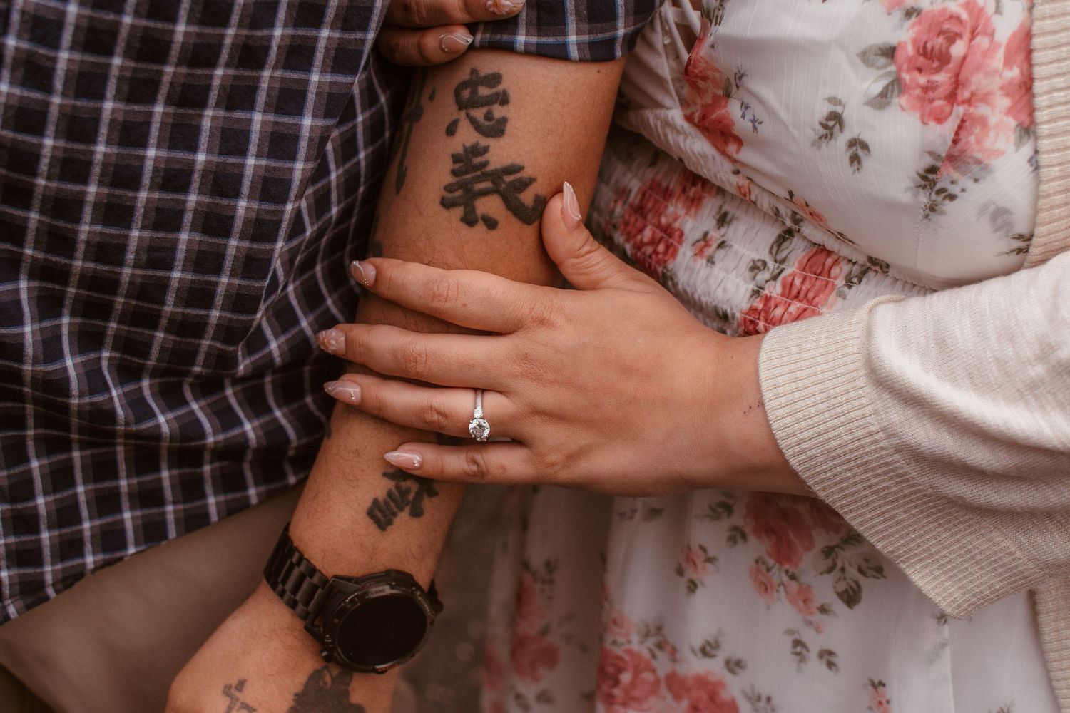 Close-up of a couple's arms intertwined, showing an engagement ring and tattoos, symbolizing love and commitment.
