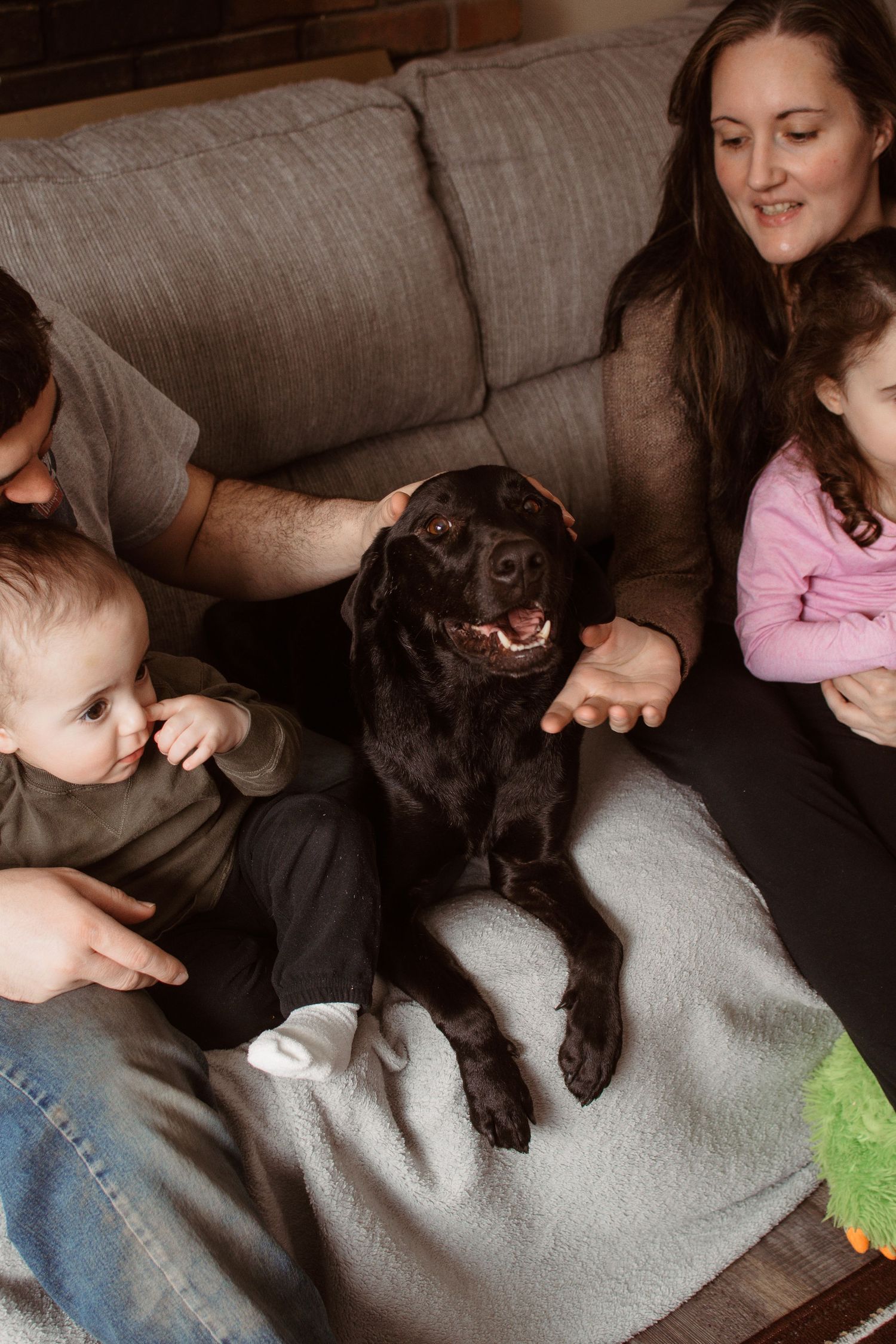 Family with toddler and young girl sitting on couch with chocolate Labrador dog on cozy blanket at home.