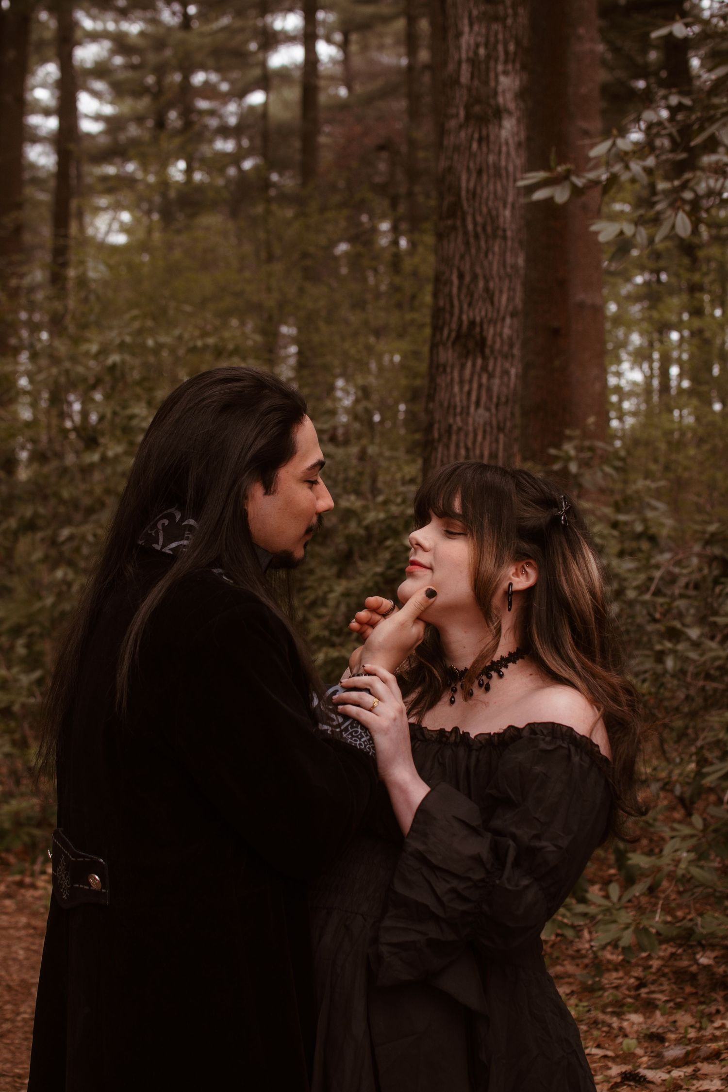 A gothic couple in black Victorian attire share a romantic moment in a misty forest setting.