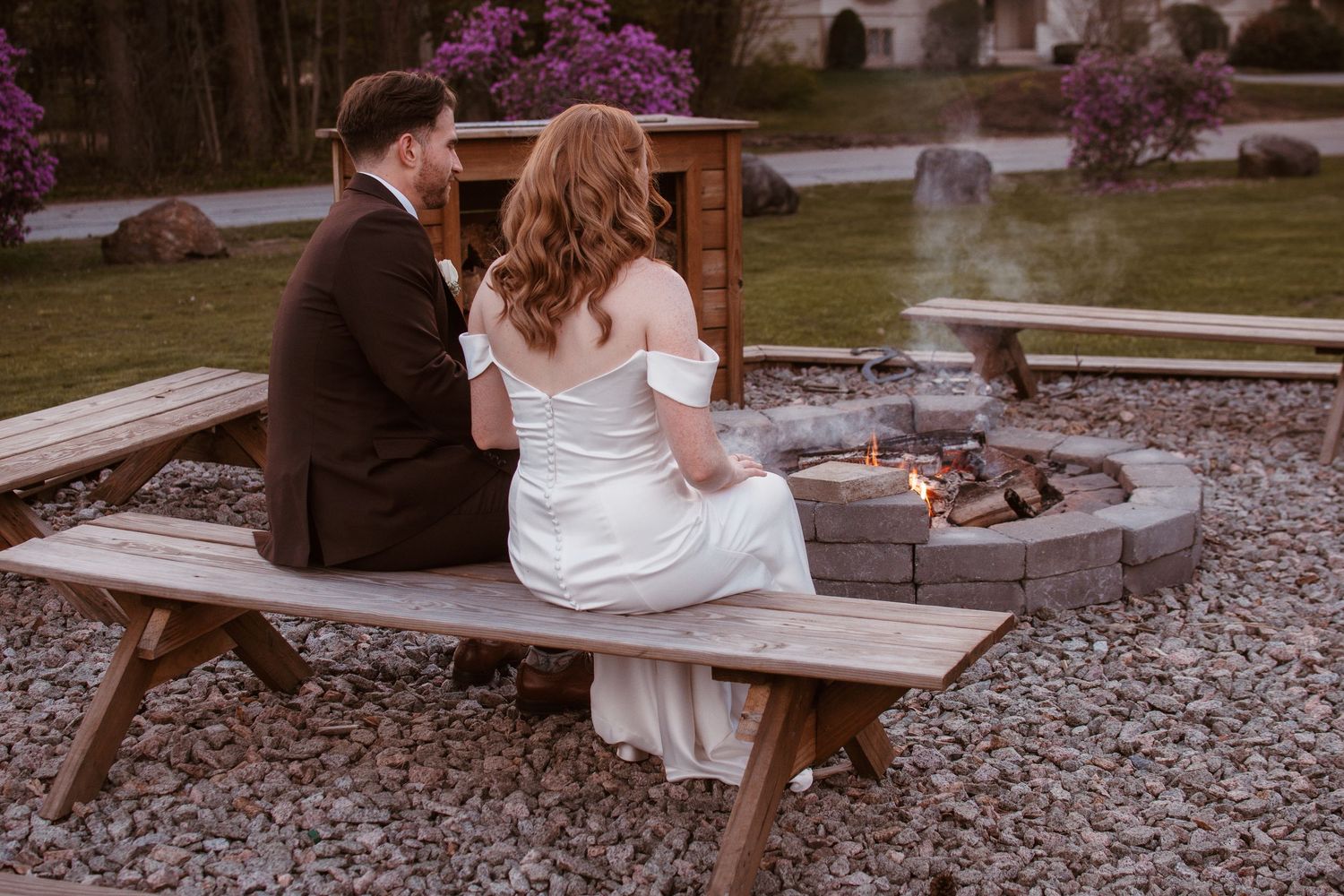Bride and groom sitting together on a wooden bench by a stone fire pit at their outdoor wedding venue.