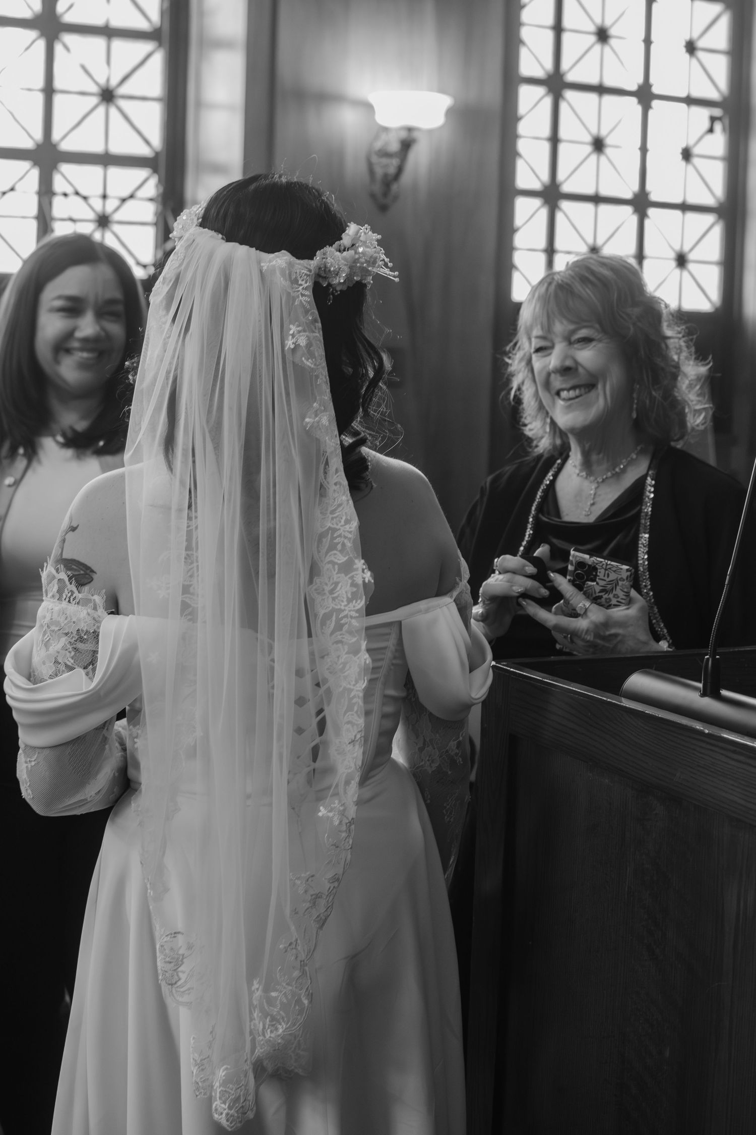 Bride in lace-trimmed veil exchanges vows with smiling officiant during wedding ceremony in black and white photo.