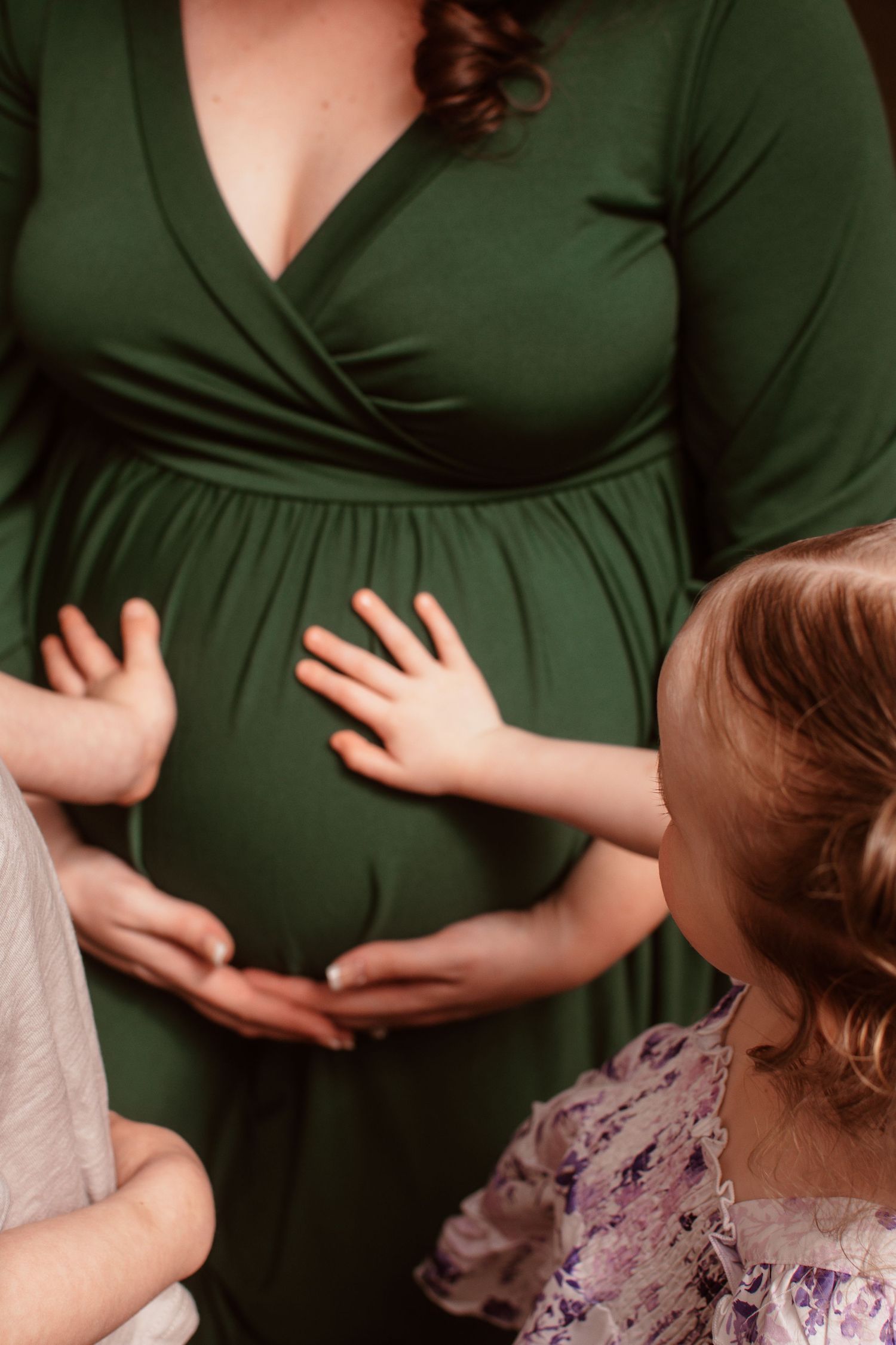 Small child in floral dress touches pregnant woman's belly while wearing a green wrap maternity dress during photo session.