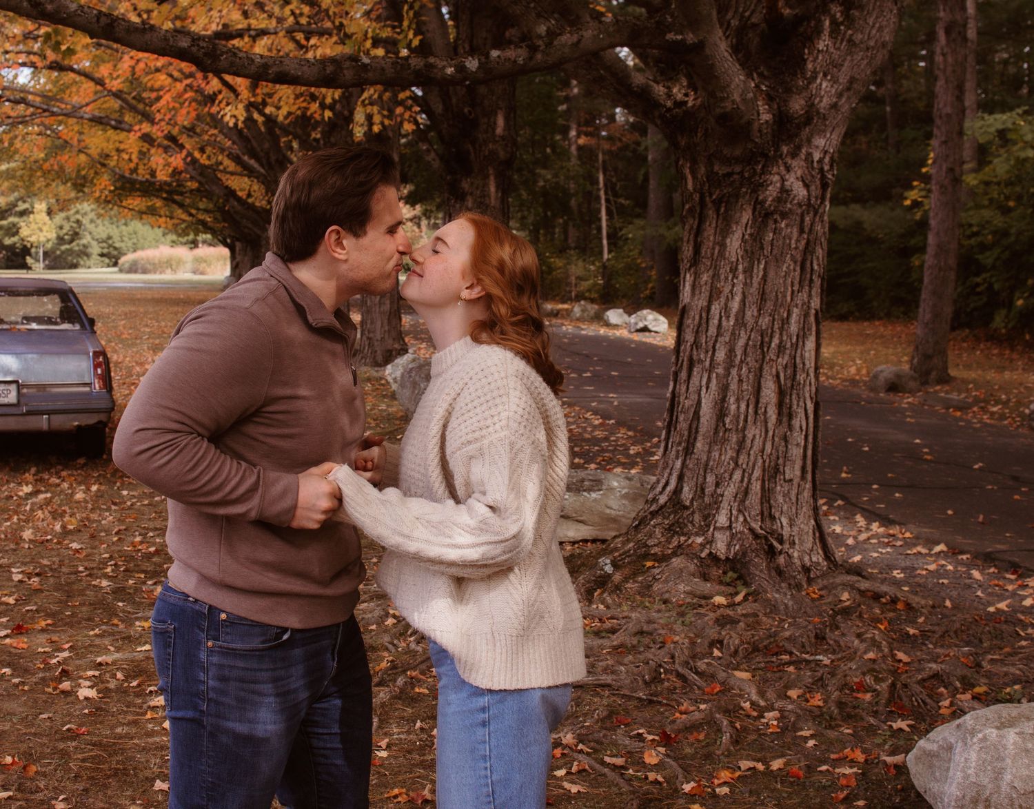 A couple shares a tender moment among fallen autumn leaves in a park setting.
