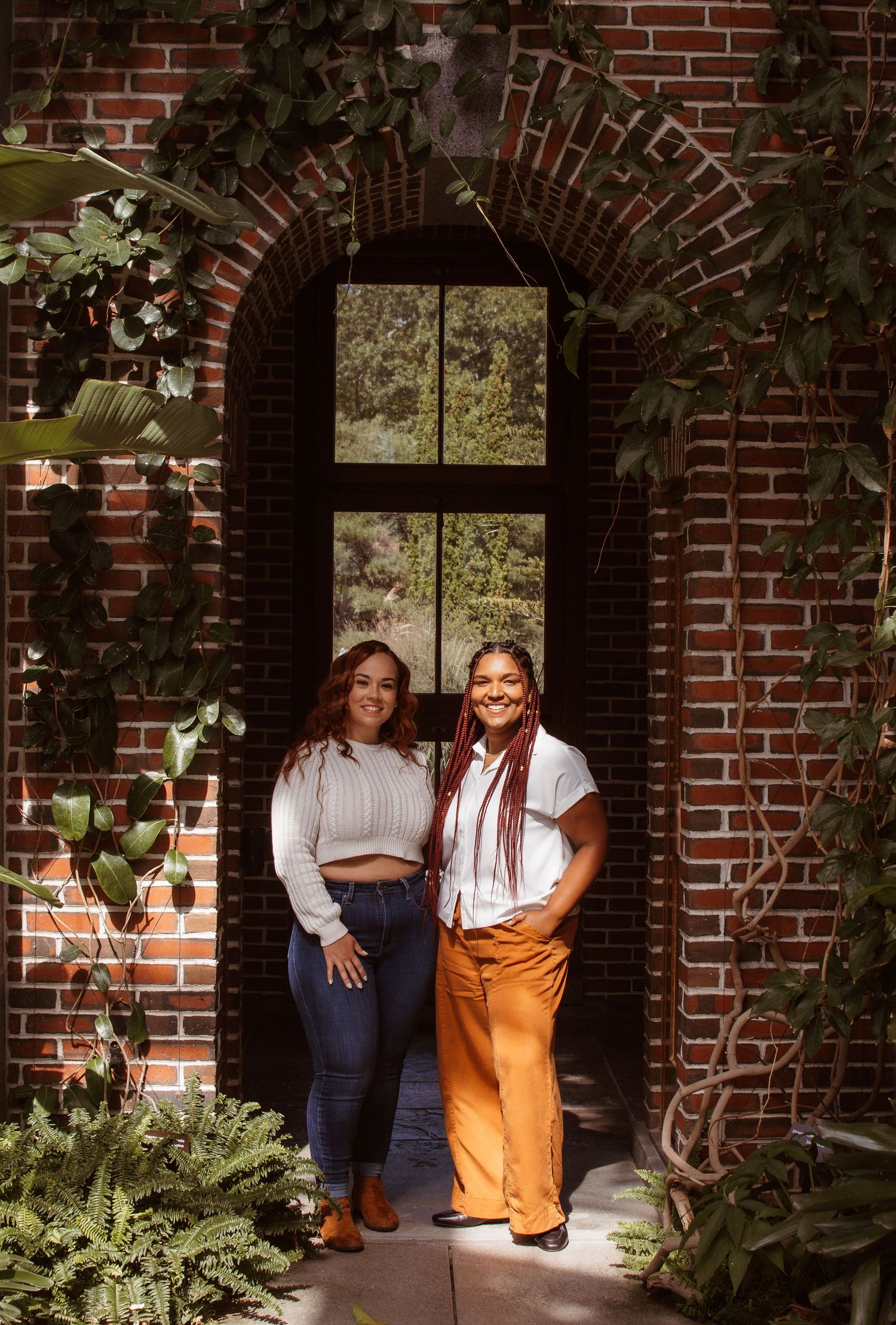 Two people stand together in an arched brick doorway surrounded by climbing vines and greenery.