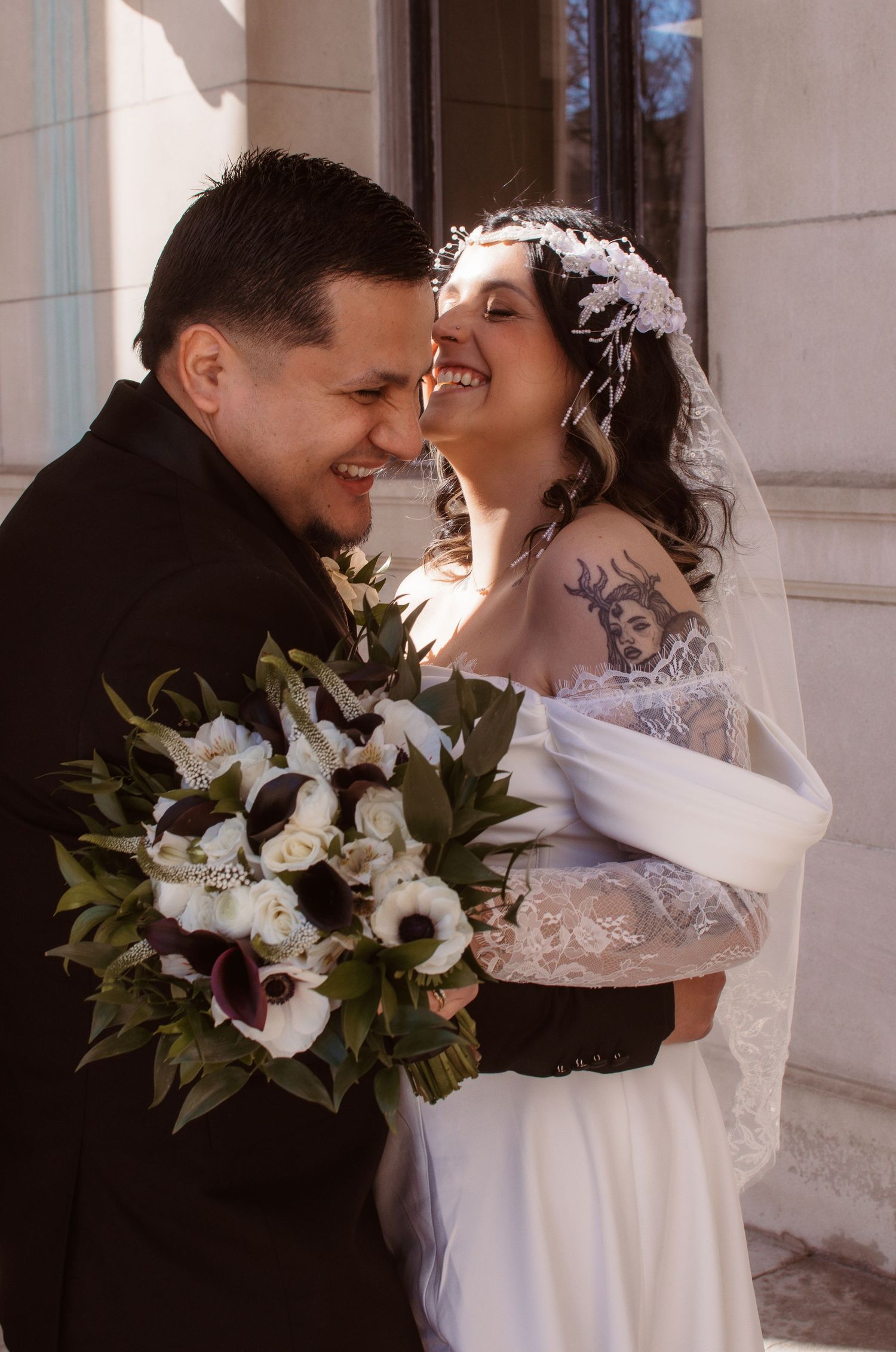 A bride and groom share a romantic moment, she wears lace gown with veil, he holds dark floral bouquet.