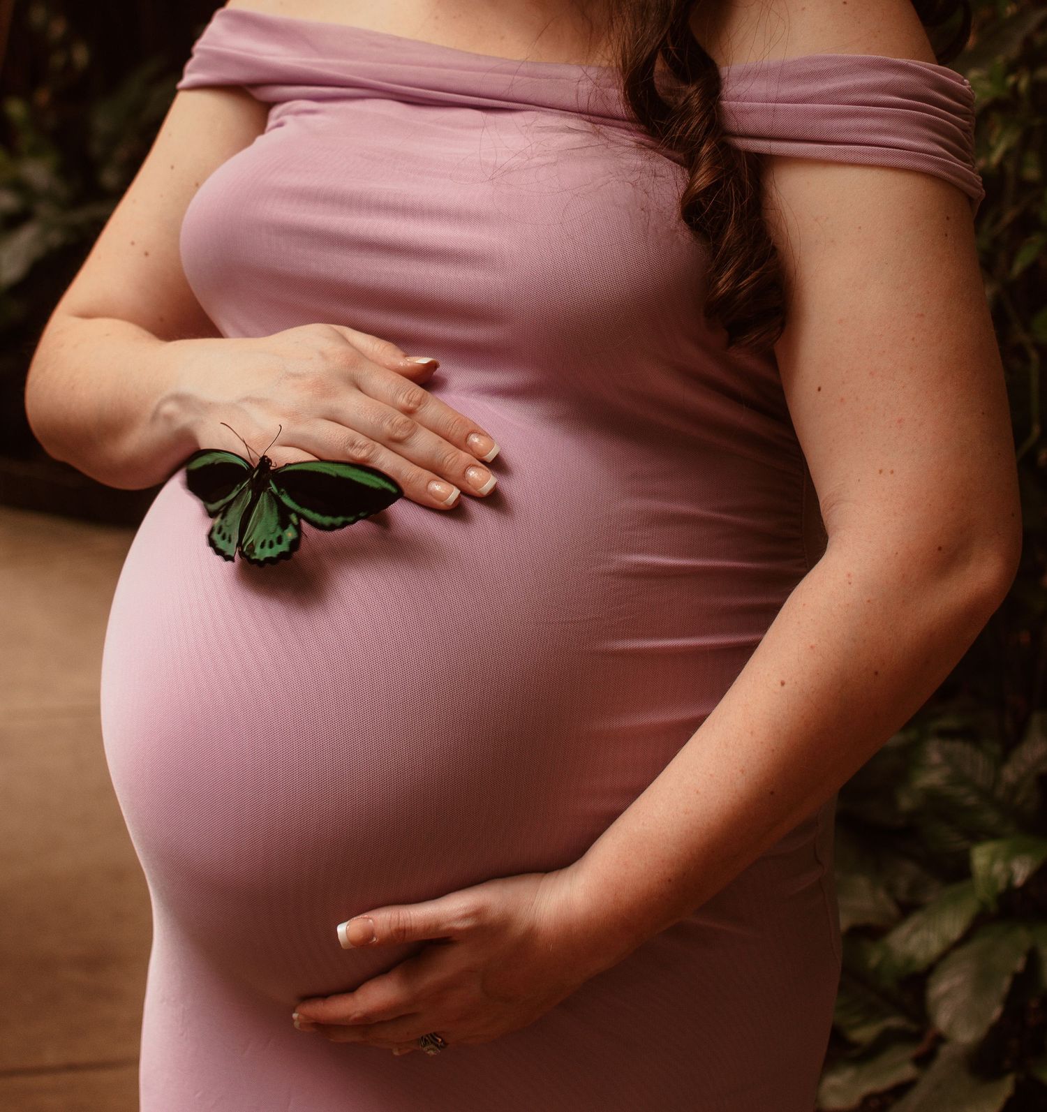 A pregnant woman in a pink dress holds a green butterfly while gently cradling her baby bump in an outdoor maternity photo.