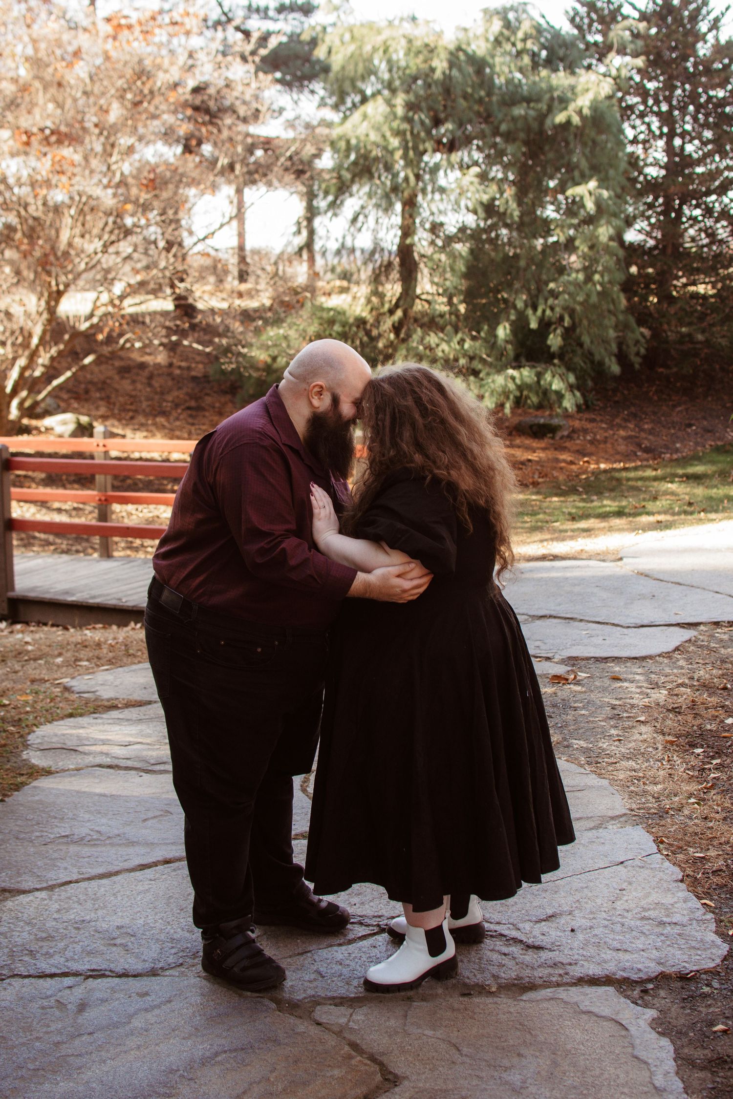 Two people in black outfits share an intimate embrace on a stone path surrounded by autumn trees in a park setting.
