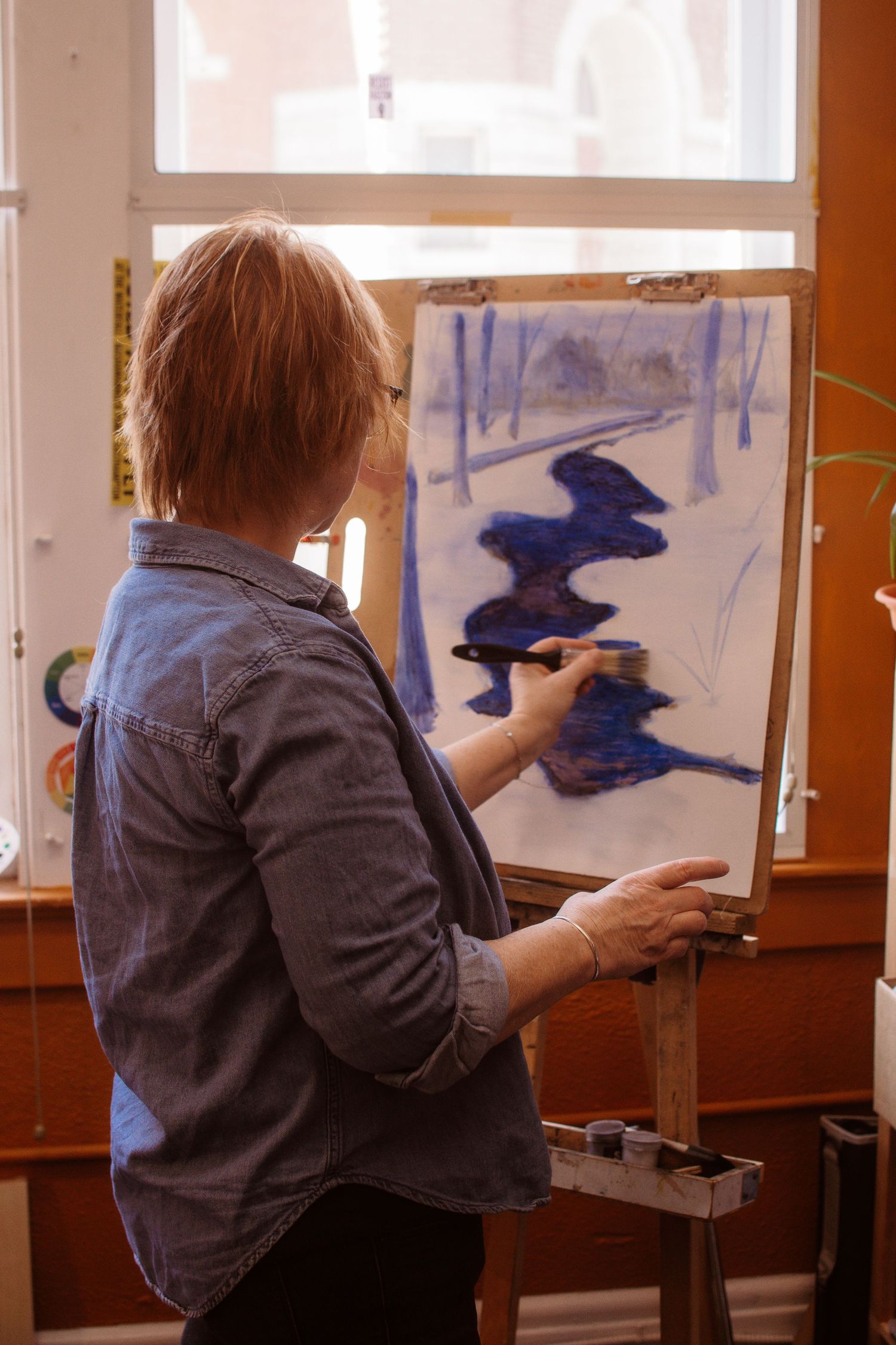 Woman painting a blue winter landscape on canvas at an easel in a bright art studio.