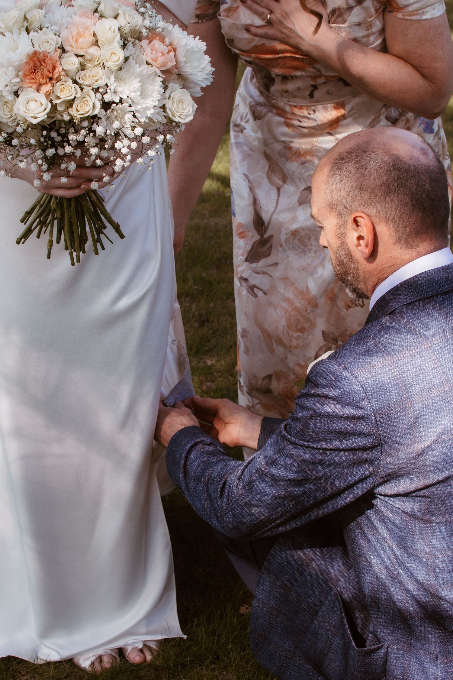 Groom kneeling to adjust bride's white wedding dress hem while she holds a peach and white rose bouquet outdoors.
