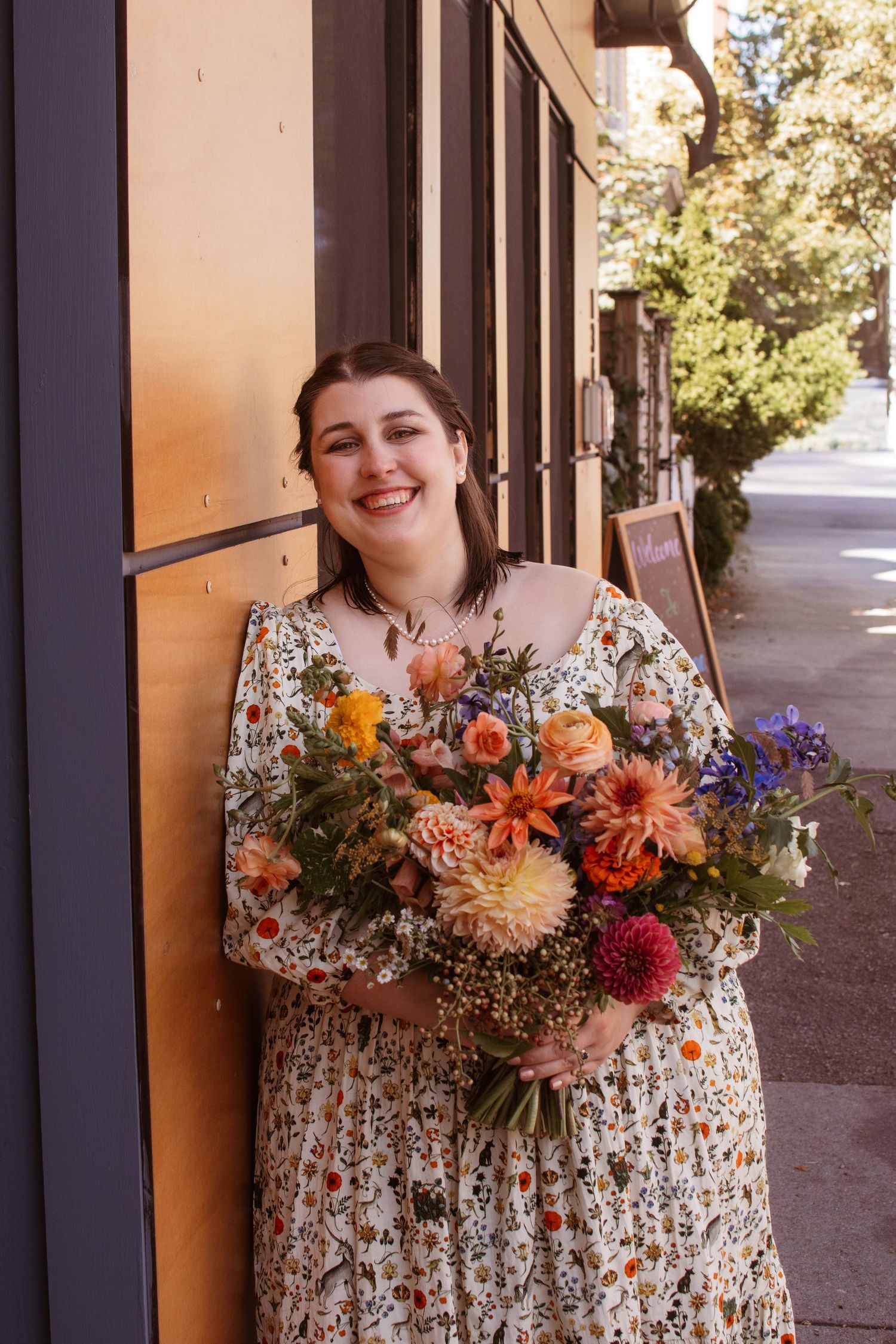 Someone wearing a floral dress holds a vibrant bouquet of dahlias and wildflowers on a sunny sidewalk.