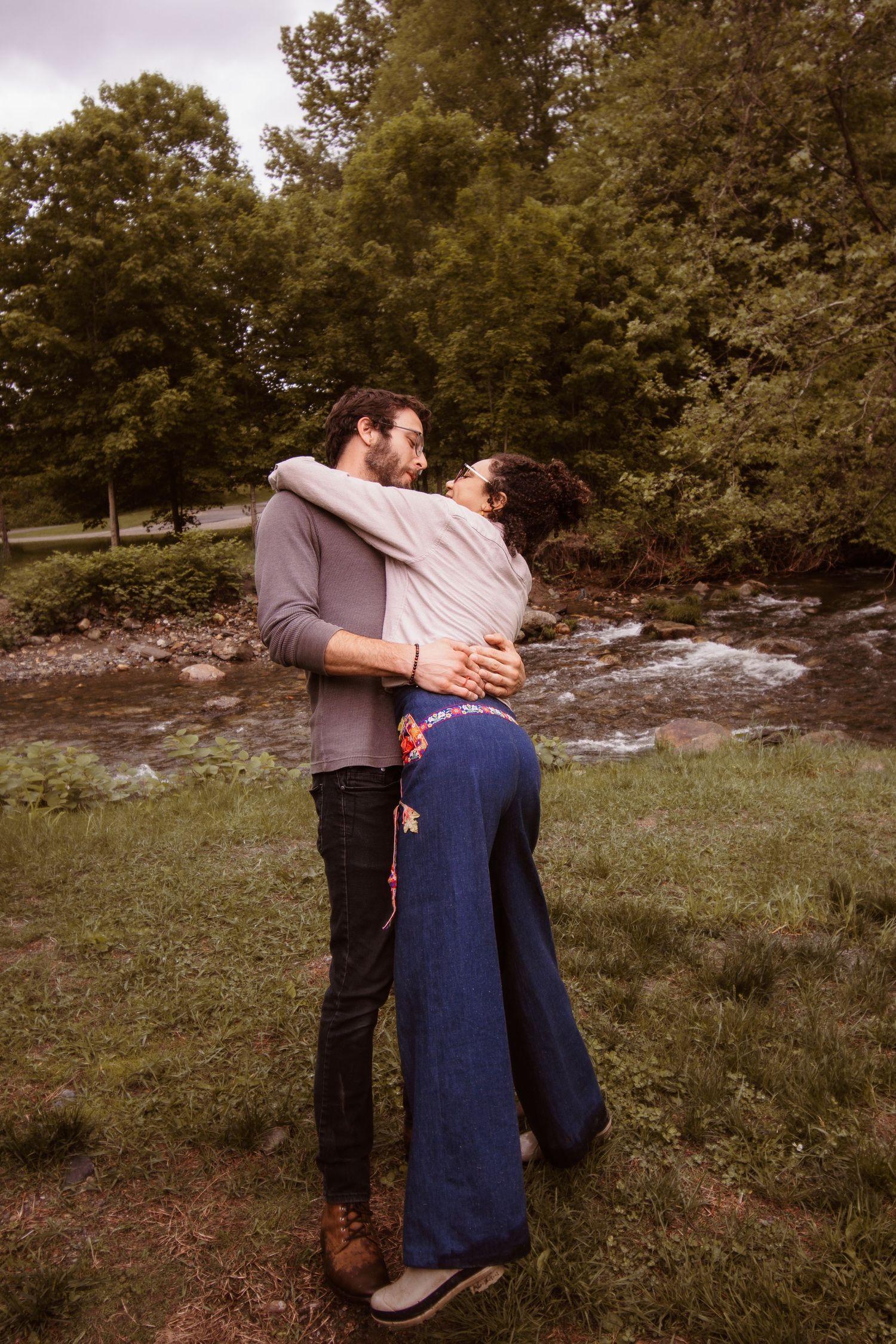 Two figures embrace in a romantic outdoor setting with trees and natural landscape in the background.