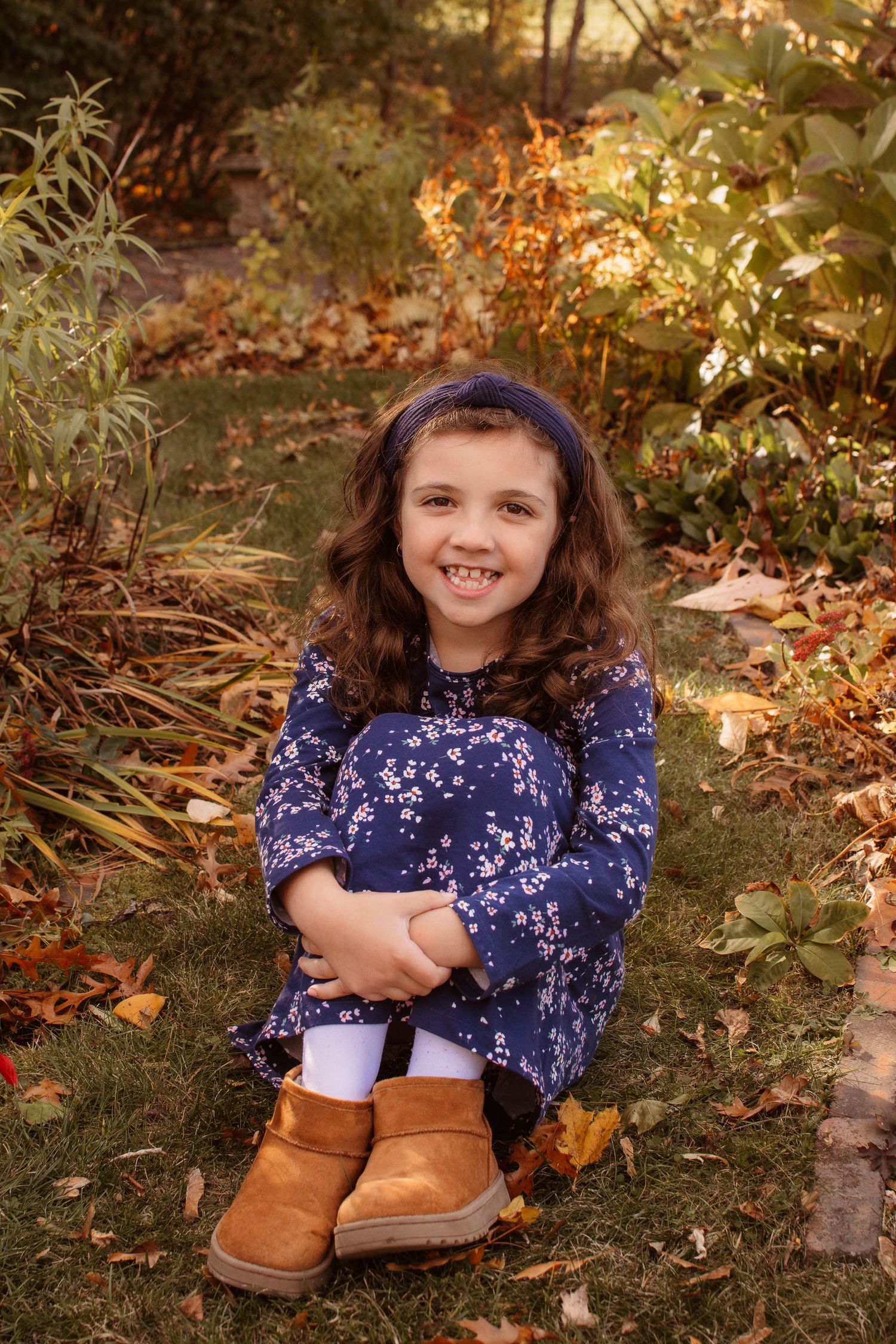 A child in a navy floral dress sits among autumn leaves wearing brown boots during a fall outdoor photoshoot.