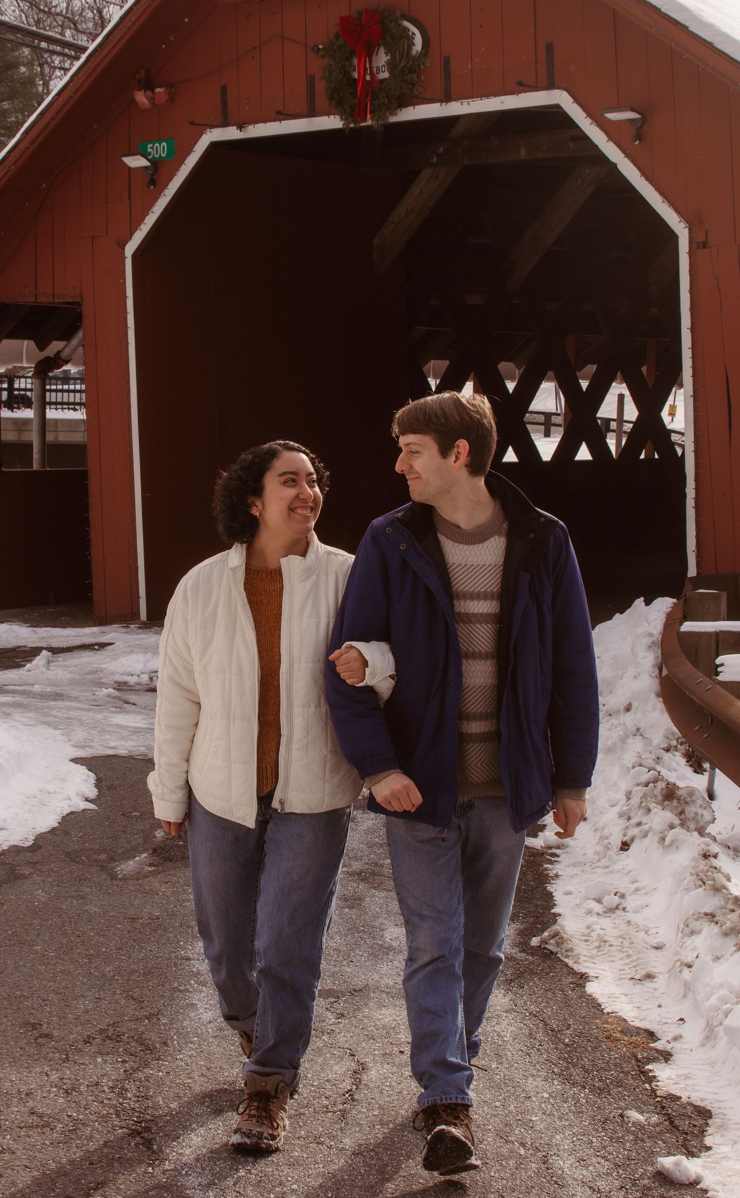 Two people walking and laughing together in winter outside a red barn with snow on the ground.