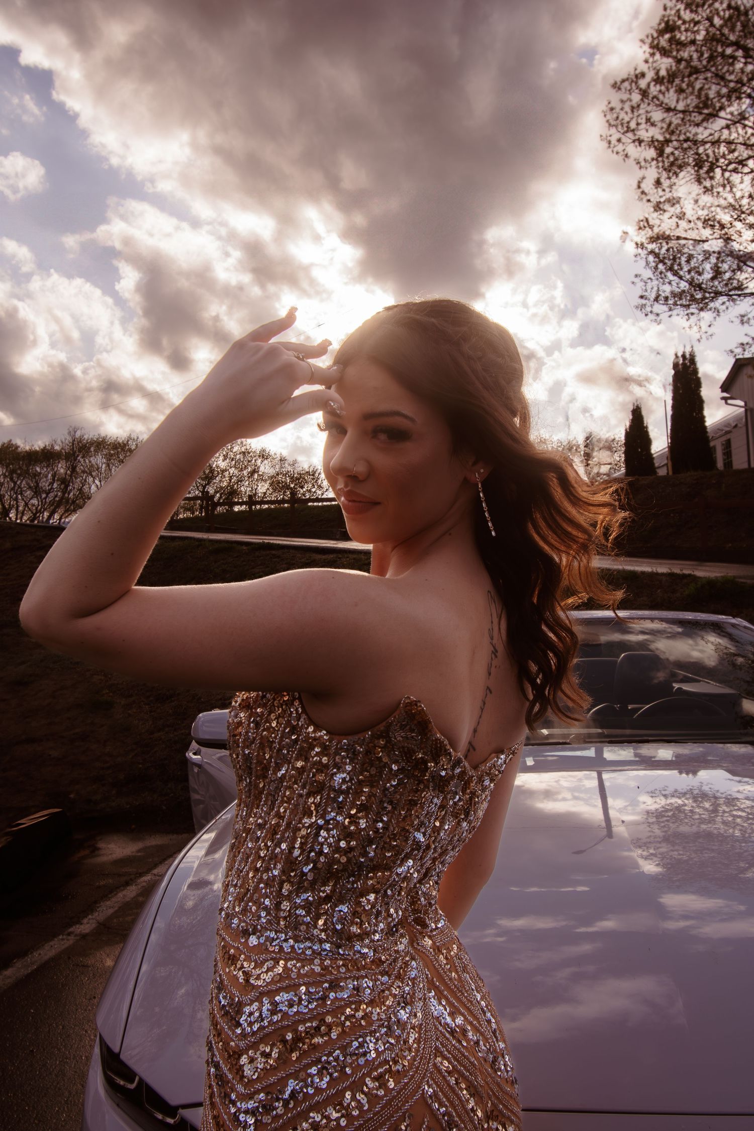 A sparkly gold sequin dress glimmers in dramatic sunset lighting next to a parked car.