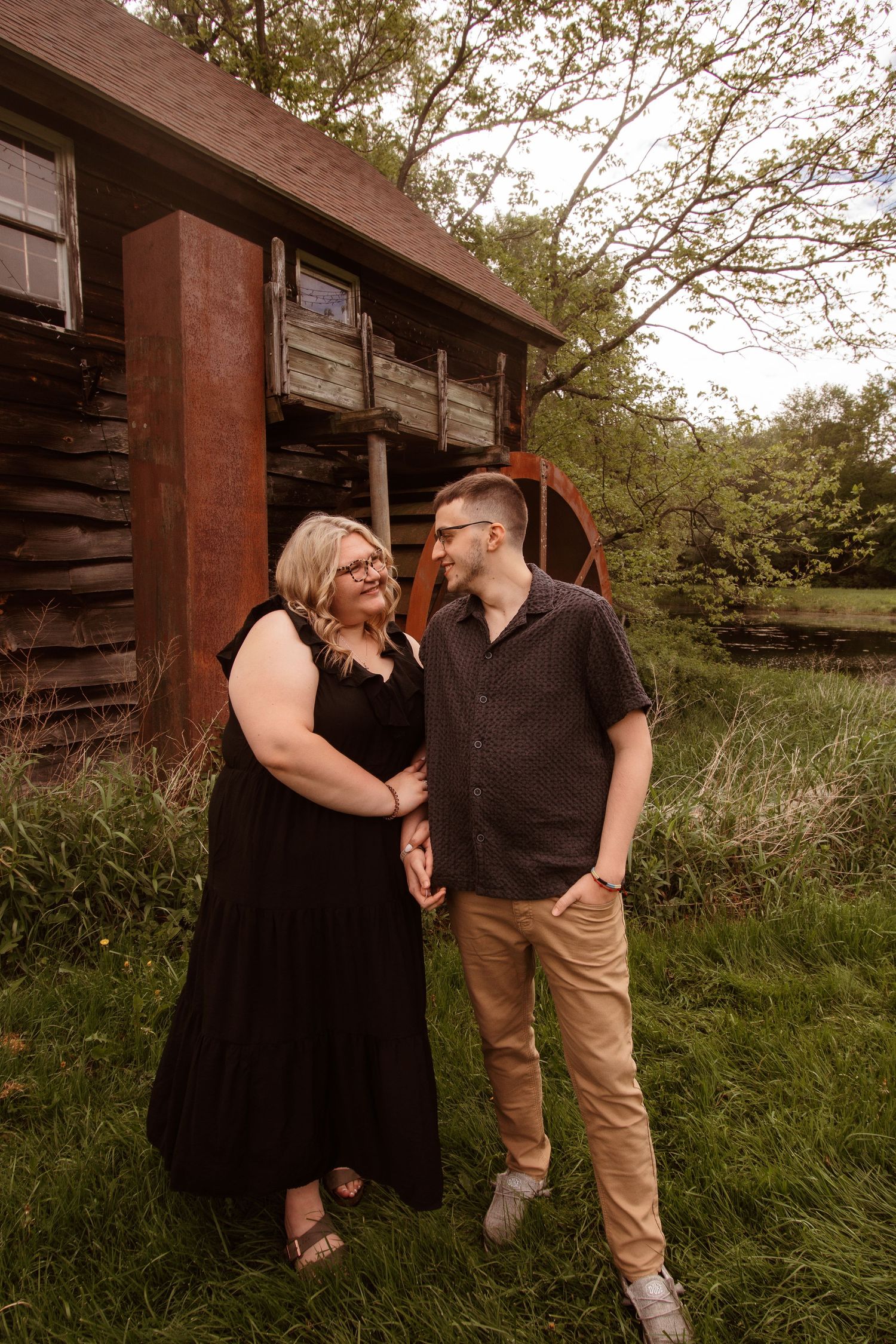 A couple shares an intimate moment while standing in tall grass beside a rustic wooden cabin at sunset.