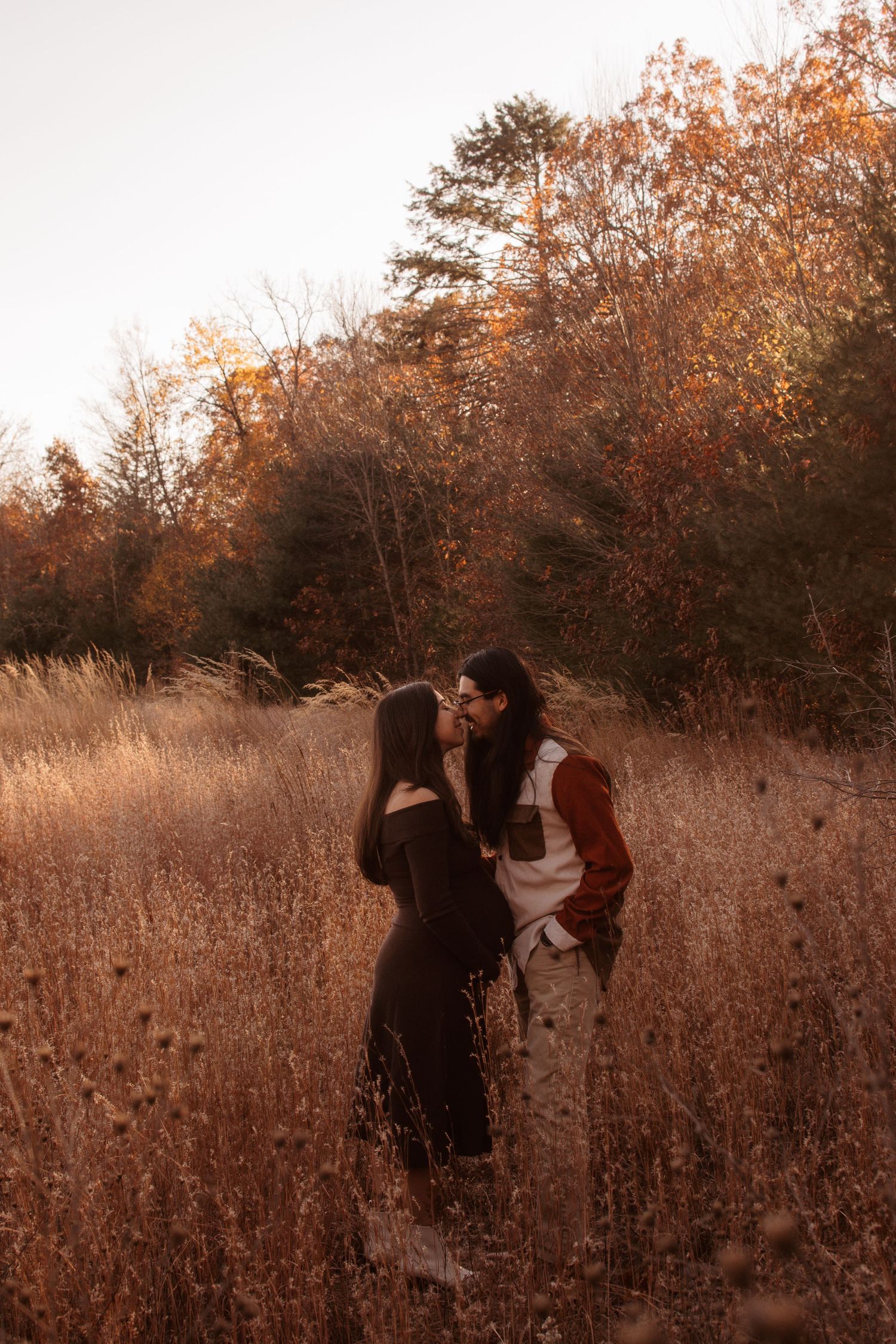 Two silhouettes embrace among tall dried grass in a golden-hued autumn field at sunset.