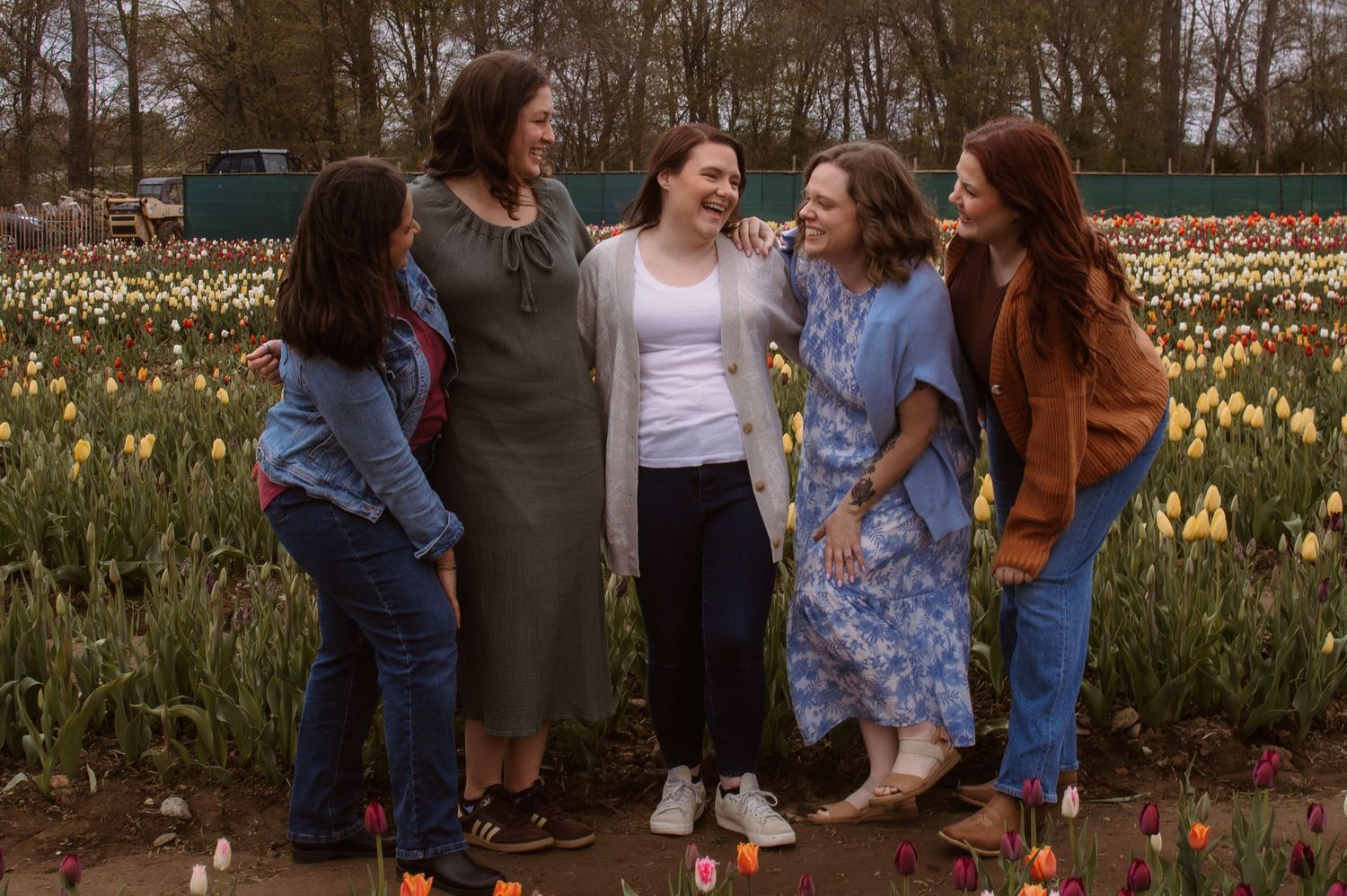 Five women laughing together in a colorful tulip field, wearing casual spring outfits in shades of blue, green, and orange.